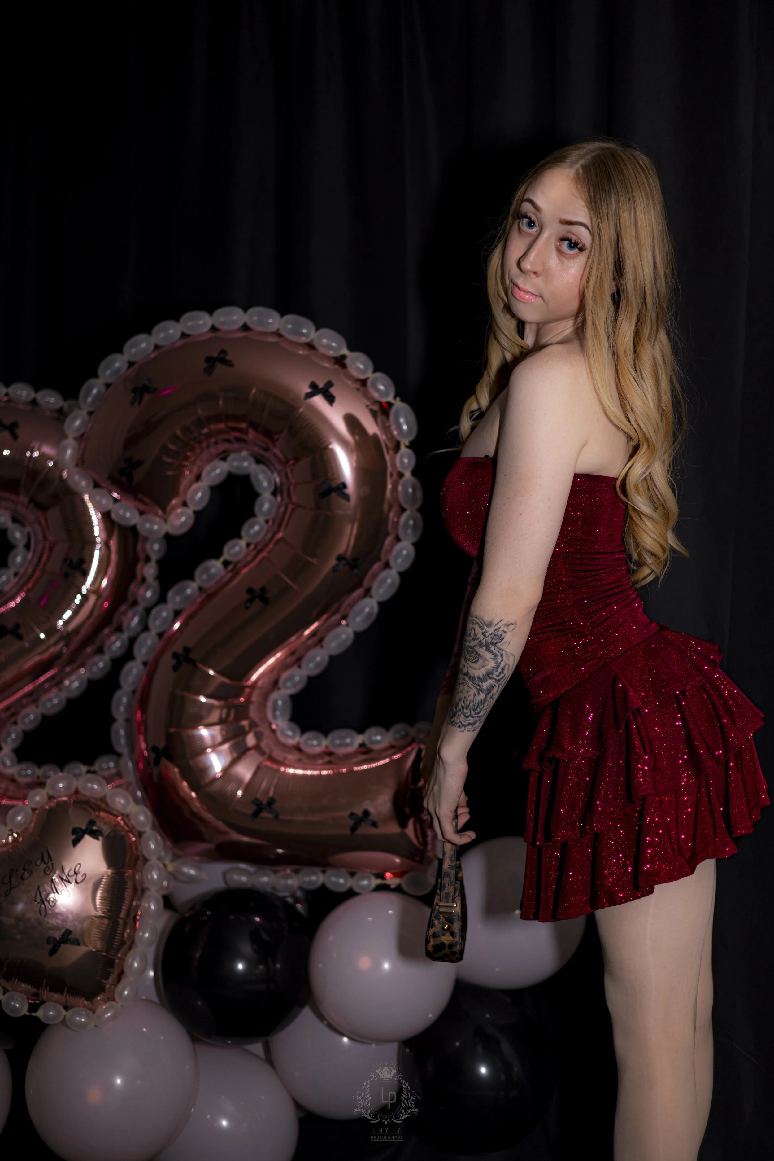 A young woman with long red hair and a tattoo on her arm, dressed in a red sparkly dress, holding a small purse, stands next to shiny balloons shaped like the number 2 and a heart at a celebration.