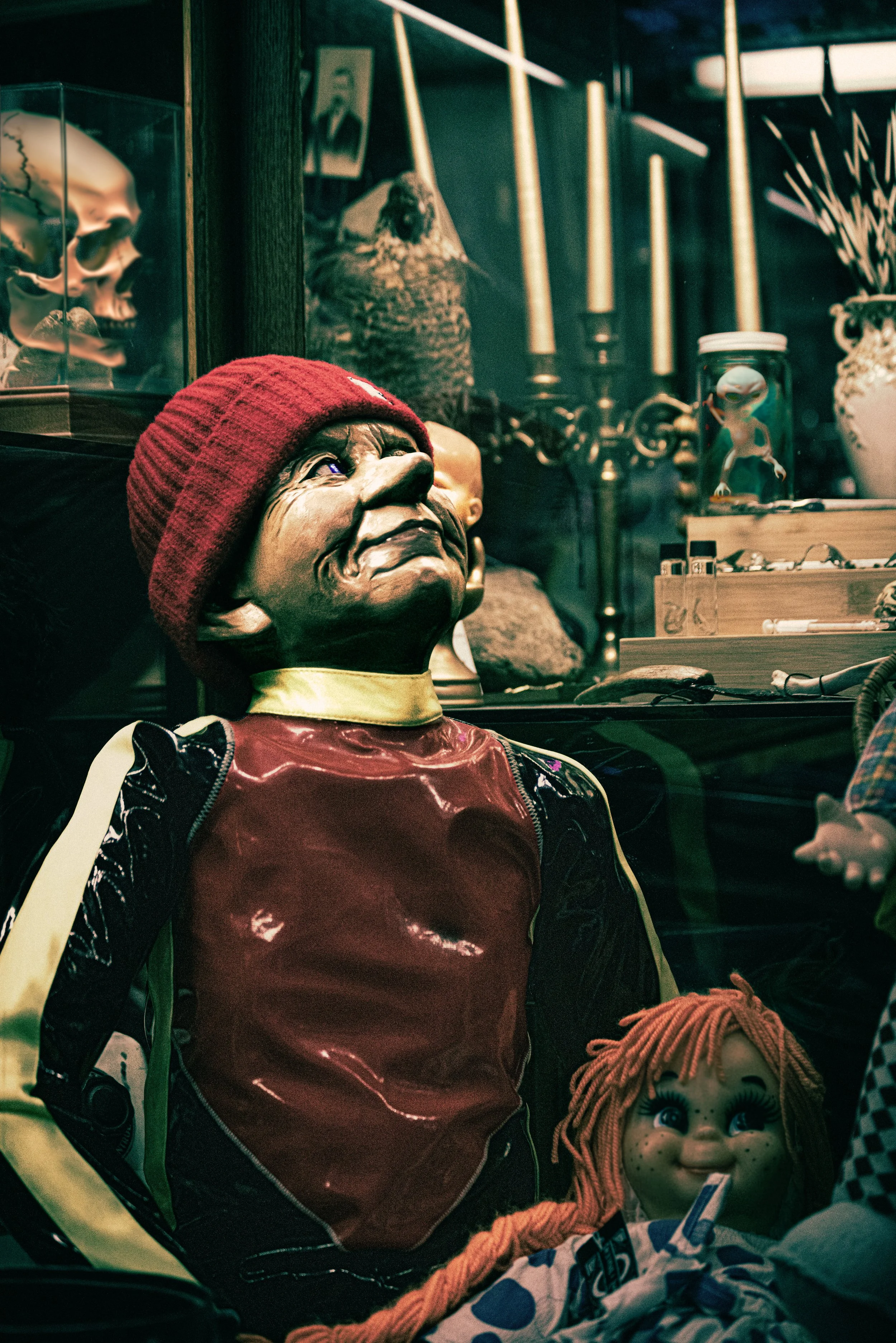 A display of creepy dolls and figurines, including a black doll with a button nose in a red beanie and shiny red jacket, an orange-haired doll with big eyes and freckles, a skull, a stuffed animal, and small figures in glass jars, set on a dark shelf with wooden and metal decorative elements.