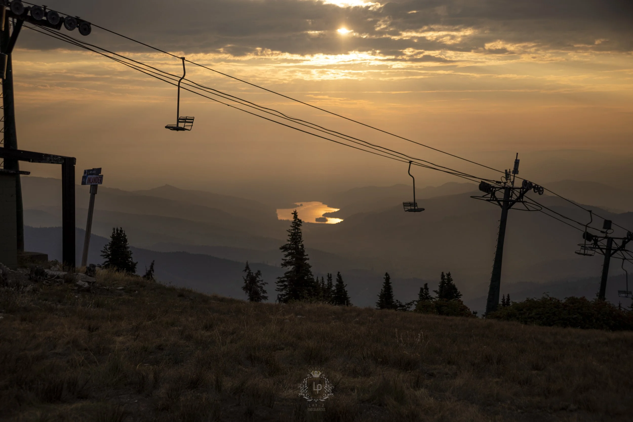 Sunset over mountainous landscape with a faint reflection on a body of water in the distance. Silhouettes of trees and ski lift chairs hanging from cables in the foreground.