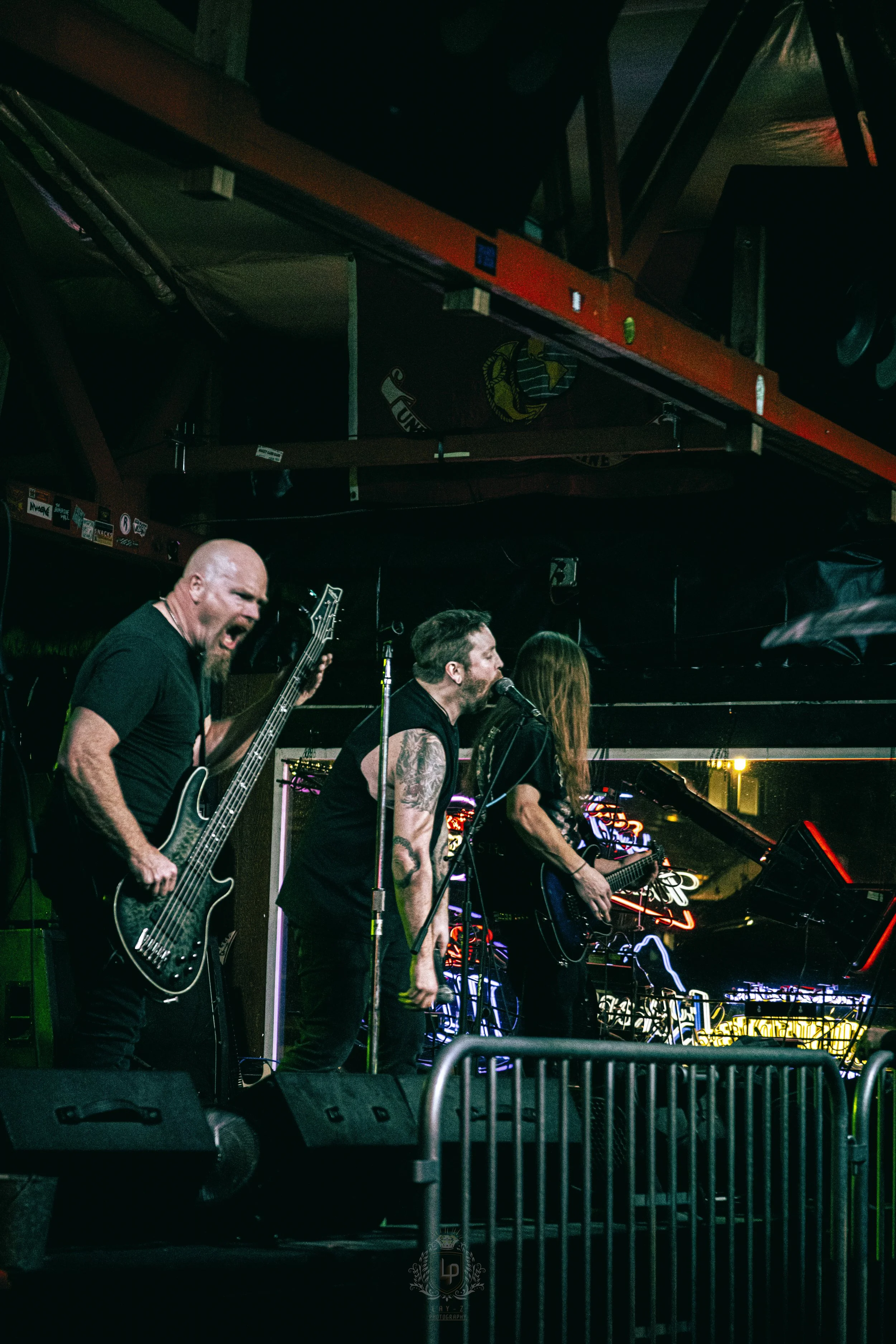 Three musicians performing on stage at a nightclub, with one playing guitar, one singing into a microphone, and another playing a guitar, illuminated by colorful neon lights.
