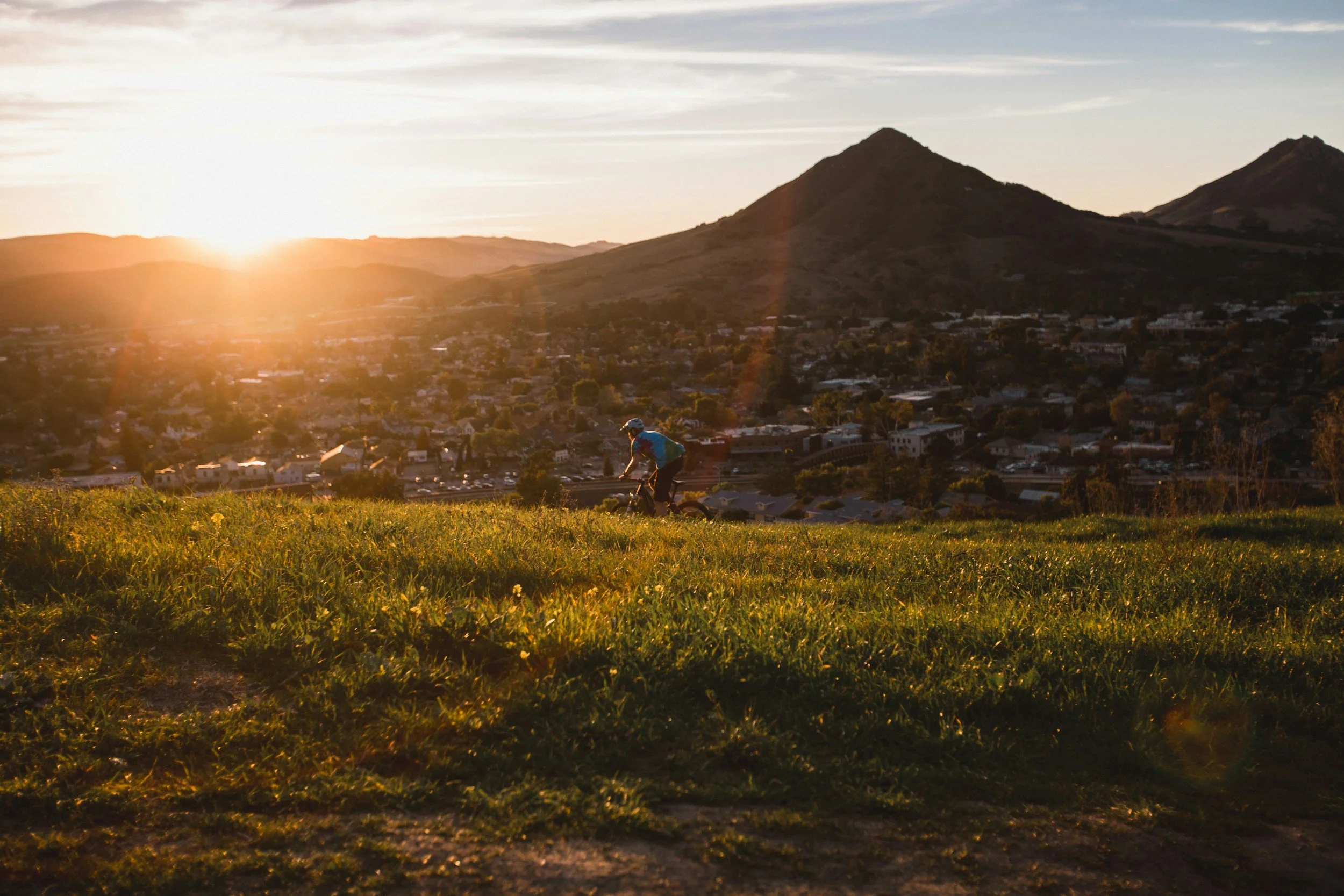 A person riding a bicycle on grassy terrain during sunset, with hills and a distant town in the background.