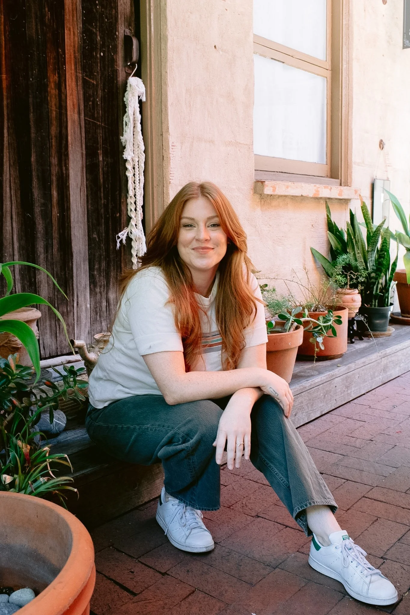 A young woman with long red hair, wearing a white t-shirt and jeans, sitting on a wooden bench outdoors, surrounded by potted plants, in front of a rustic wall with a window.