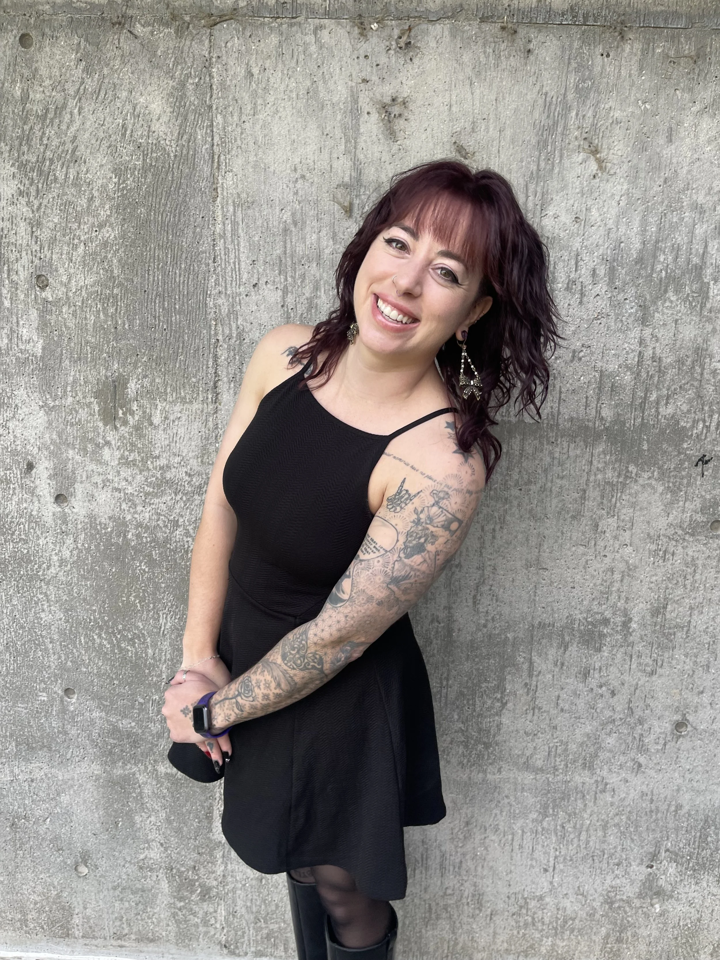 A therapist, Adare Toral, with purple hair, tattoos on her arms, and wearing a black dress, standing against a concrete wall, smiling at the camera.