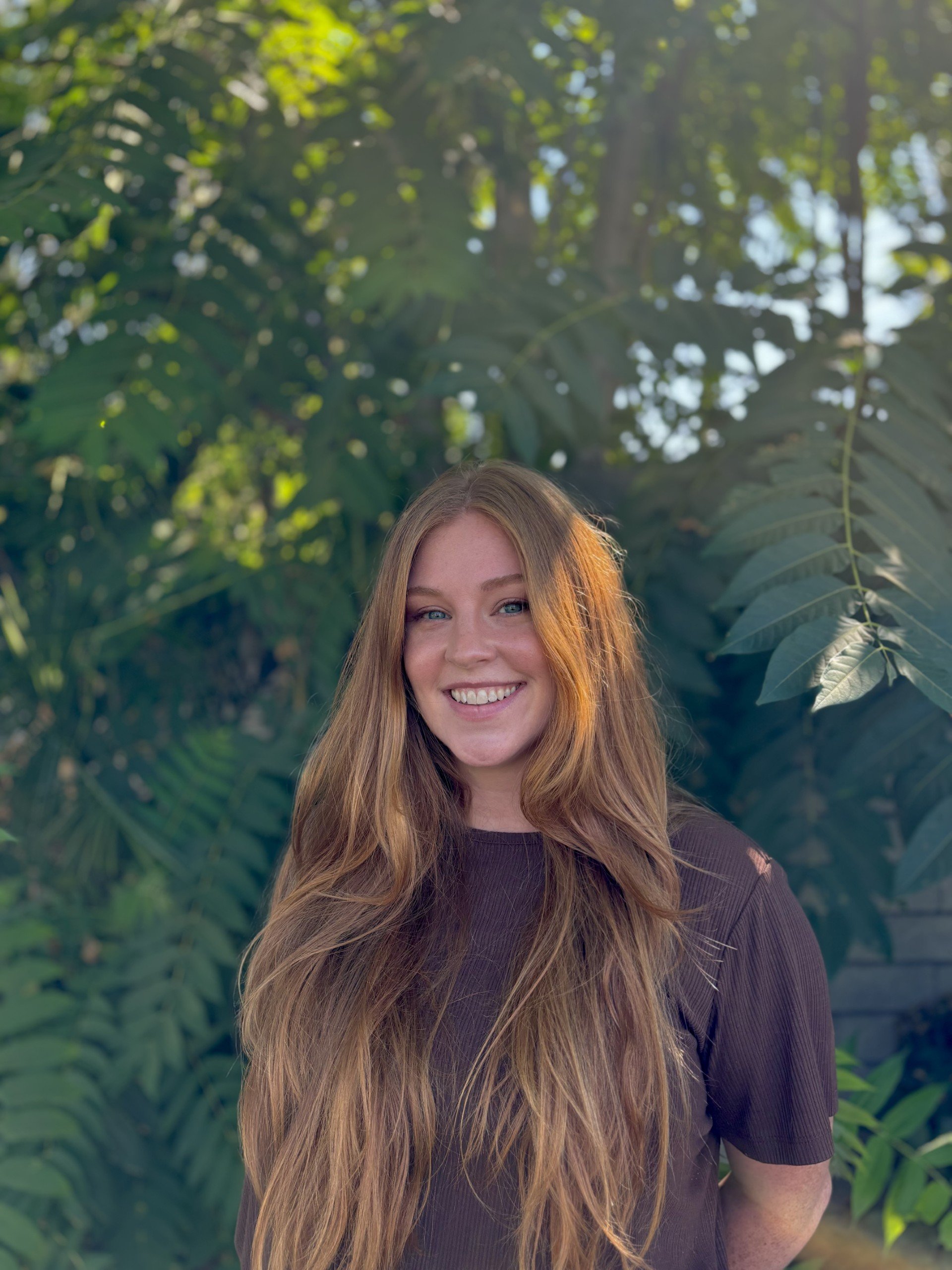 A therapist, Mary Kate Hoag, with long red hair standing outdoors in front of lush green foliage.