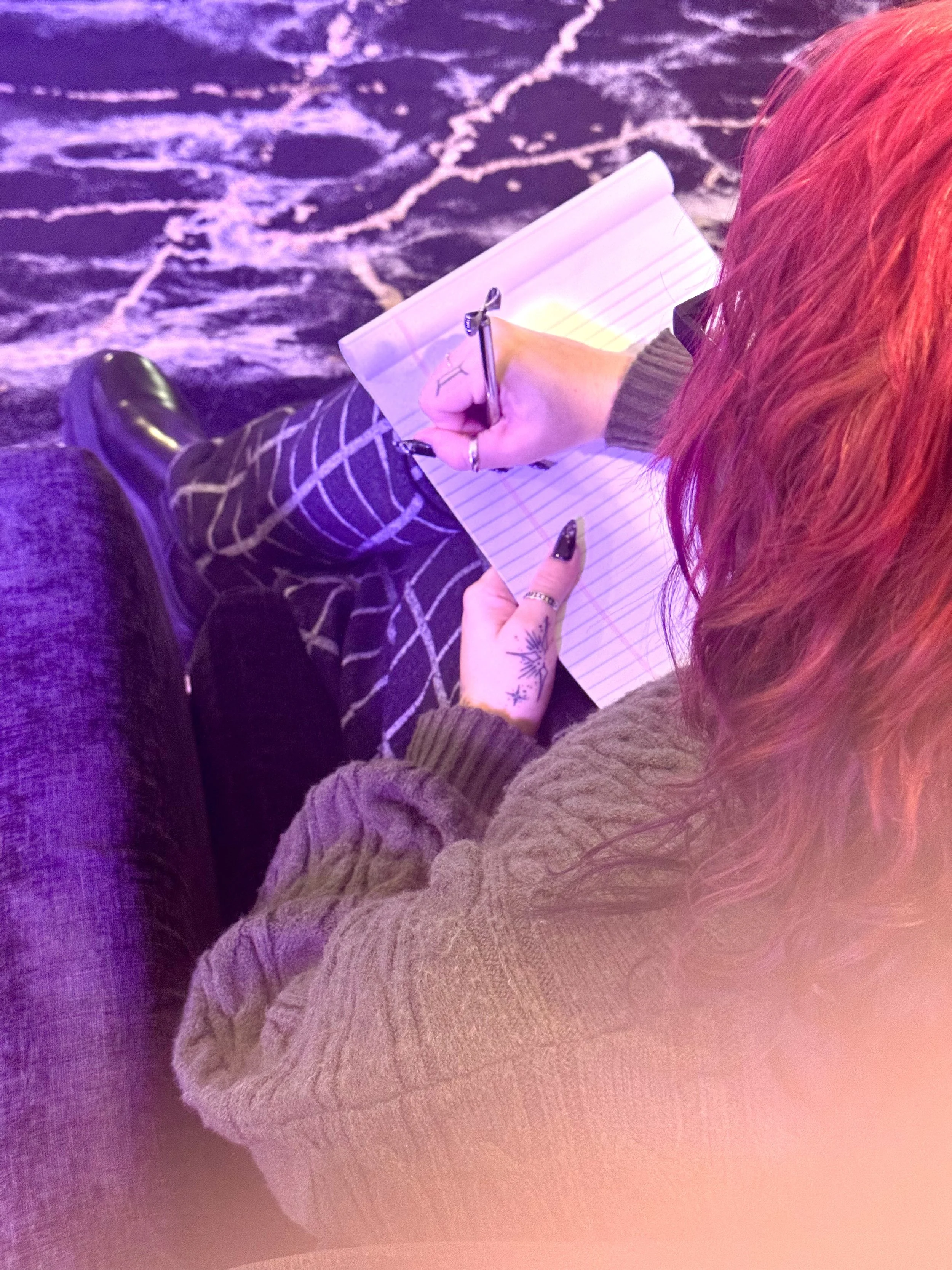 A woman with red hair writing in a notebook while sitting on a black chair on a marble patterned rug..