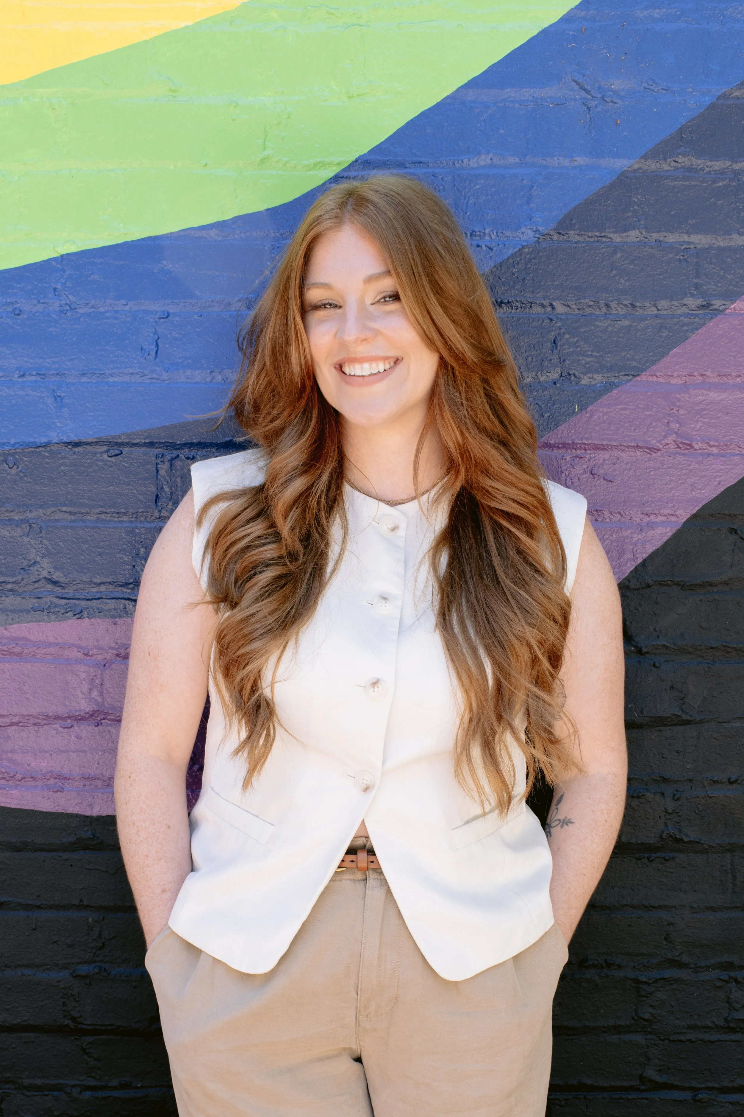 A woman with long, wavy reddish-brown hair smiling while standing in front of a colorful, rainbow-painted brick wall wearing a sleeveless white button-up vest and beige pants.