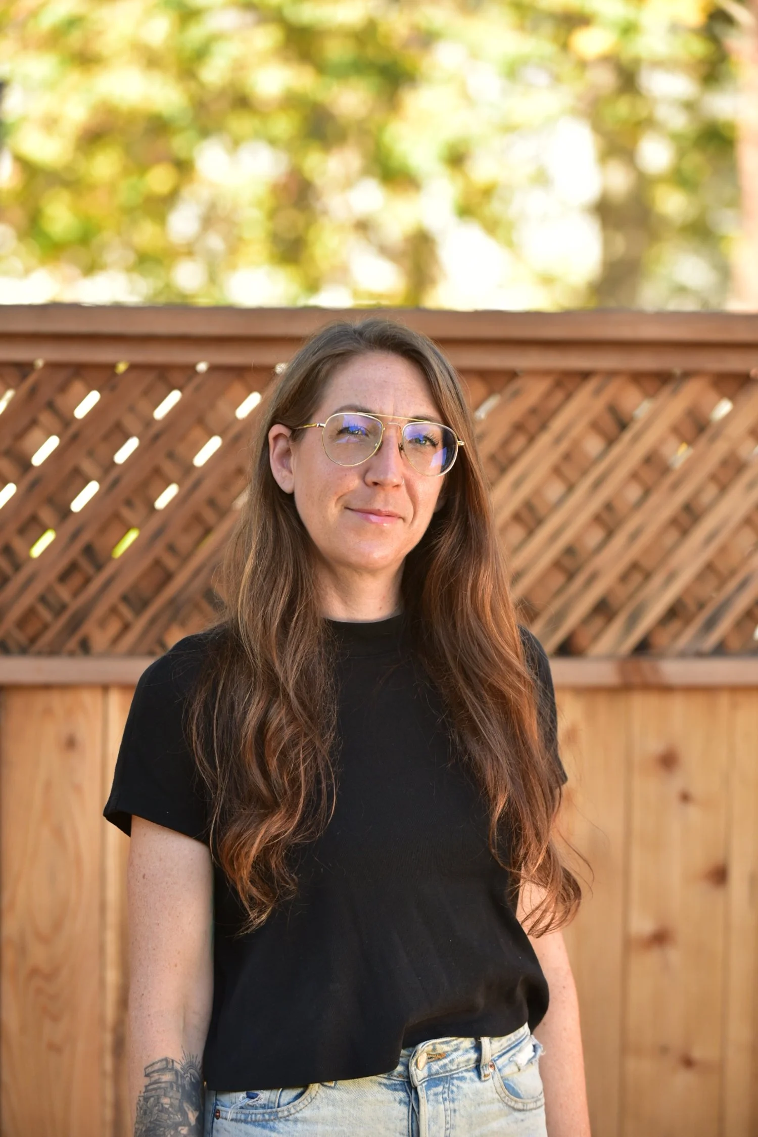A therapist, Kelly Knox, with long wavy hair and glasses standing outdoors in front of a wooden fence with trees in the background.