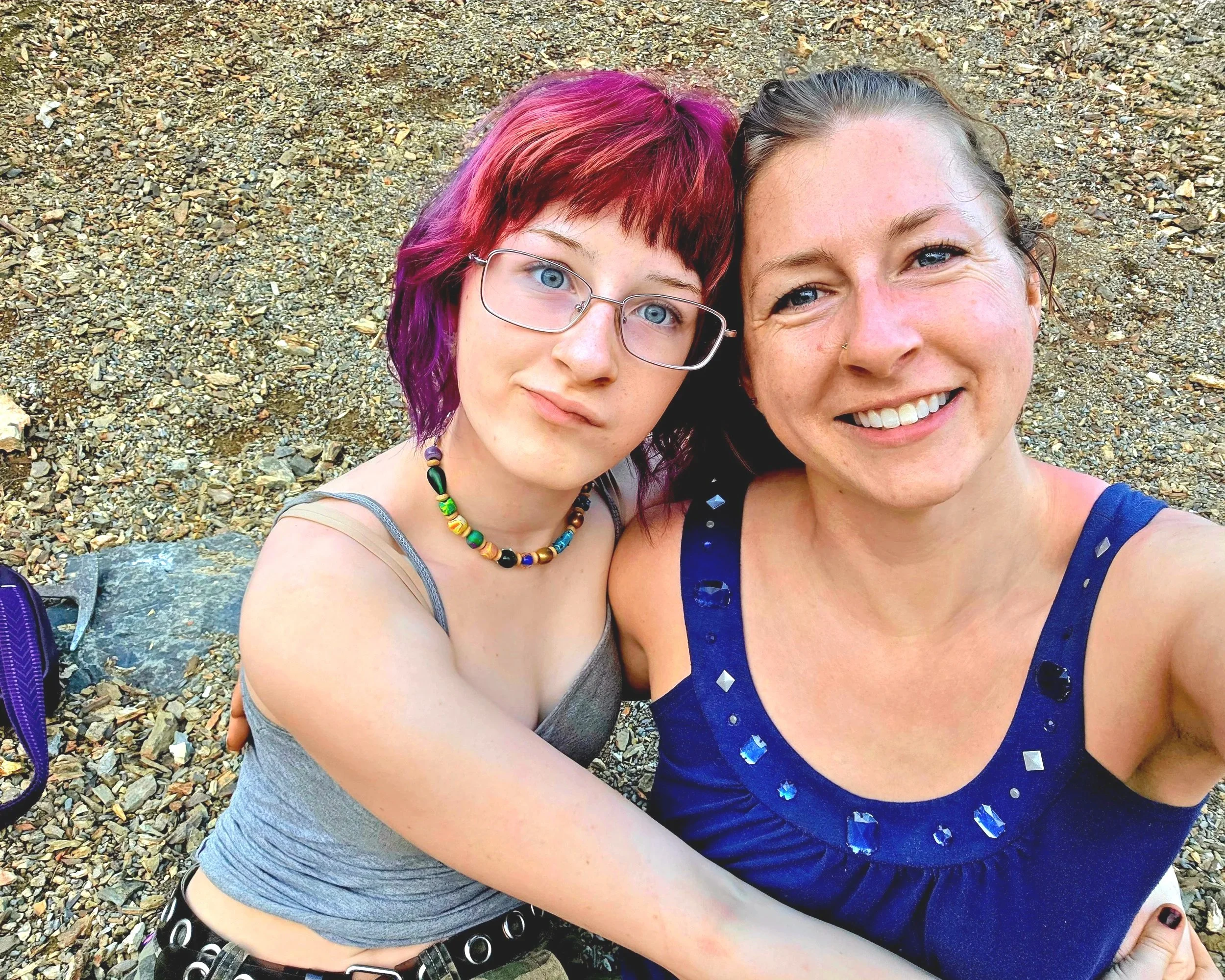 Close-up of two women taking a selfie on a rocky beach. One woman has vibrant pink and purple hair, glasses, and a colorful beaded necklace. The other woman has light brown or blonde hair tied back, freckles, and is wearing a blue dress with rhinestone embellishments. They are smiling and appear to be enjoying a sunny day.