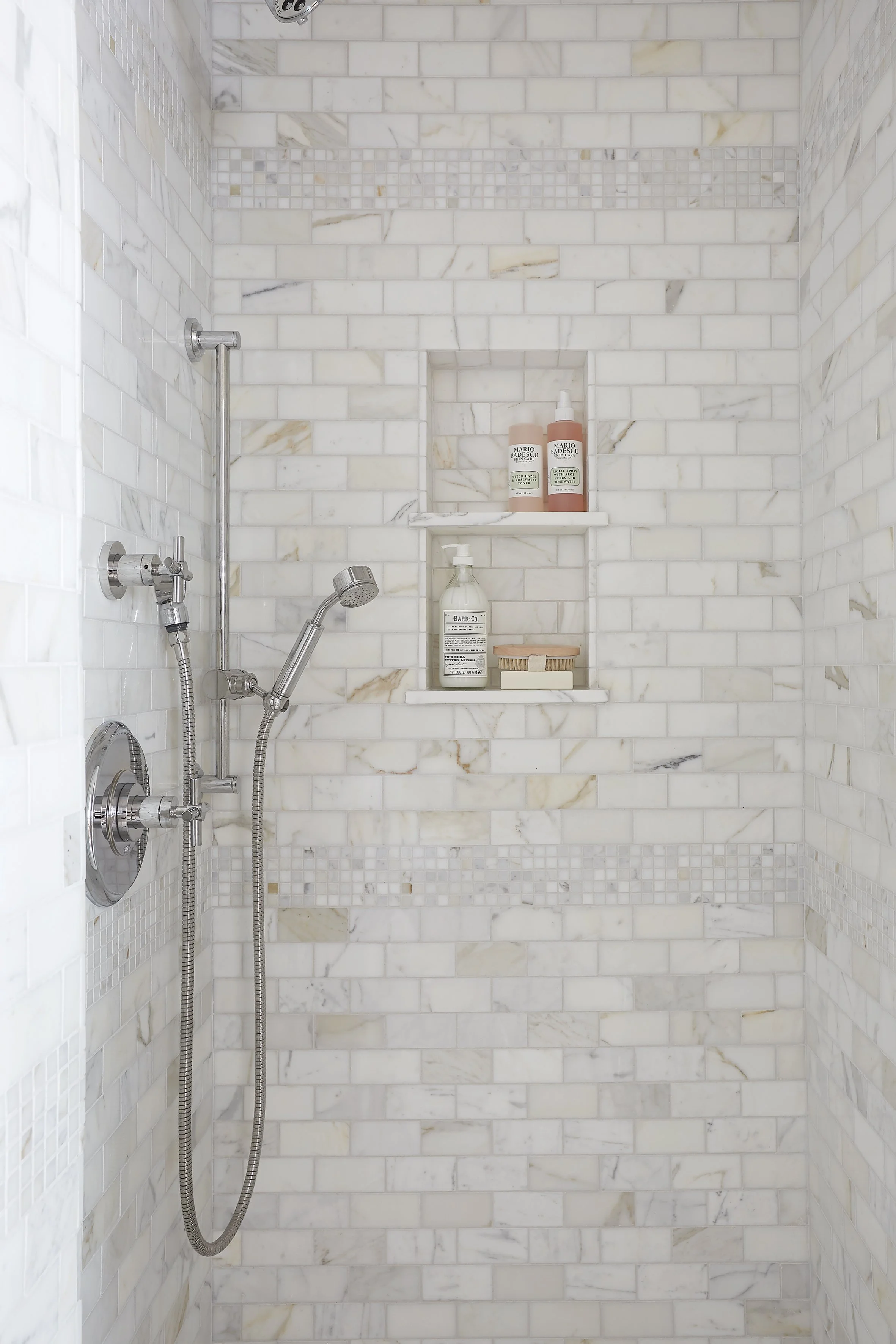 A marble shower with built-in wall niches holding shampoo, conditioner, body wash, and a scrub brush.