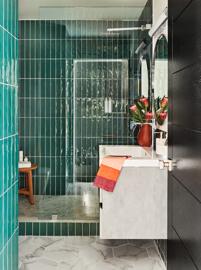Modern bathroom with teal tile walls, a shower with glass door, a concrete vanity with a white sink, a floral arrangement in a brown vase, and hexagonal marble floor tiles.