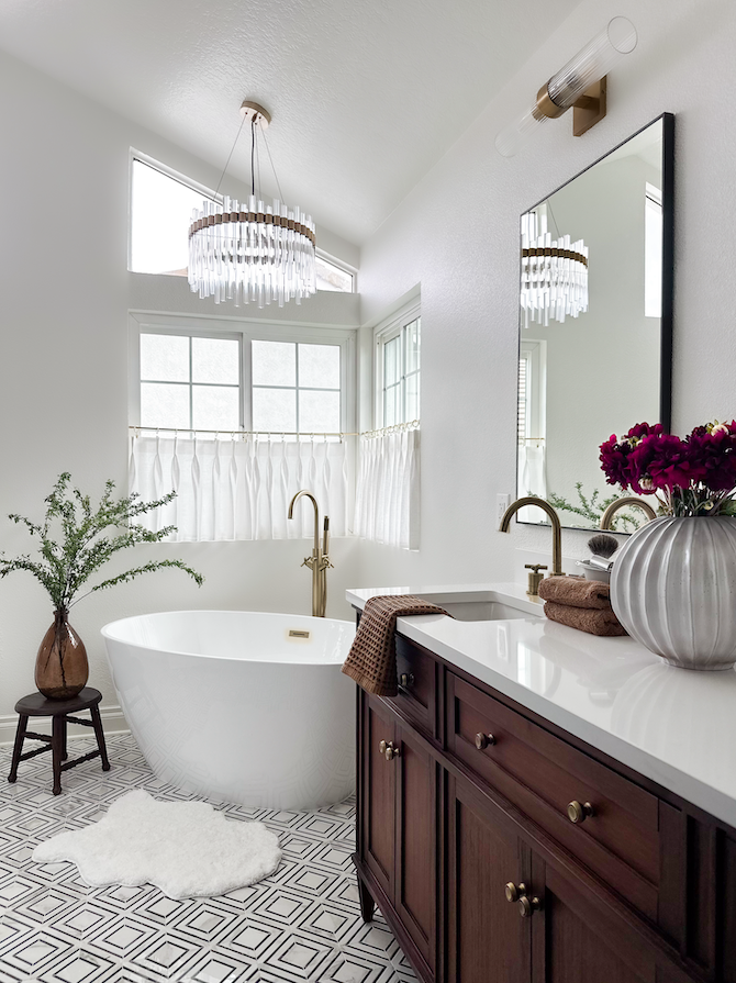 Bright bathroom with a white freestanding bathtub, a large mirror, and a dark wood vanity with a white countertop. There is a vase with pink flowers on the vanity, a potted plant on a small stool, and geometric tile flooring. A chandelier and a wall 