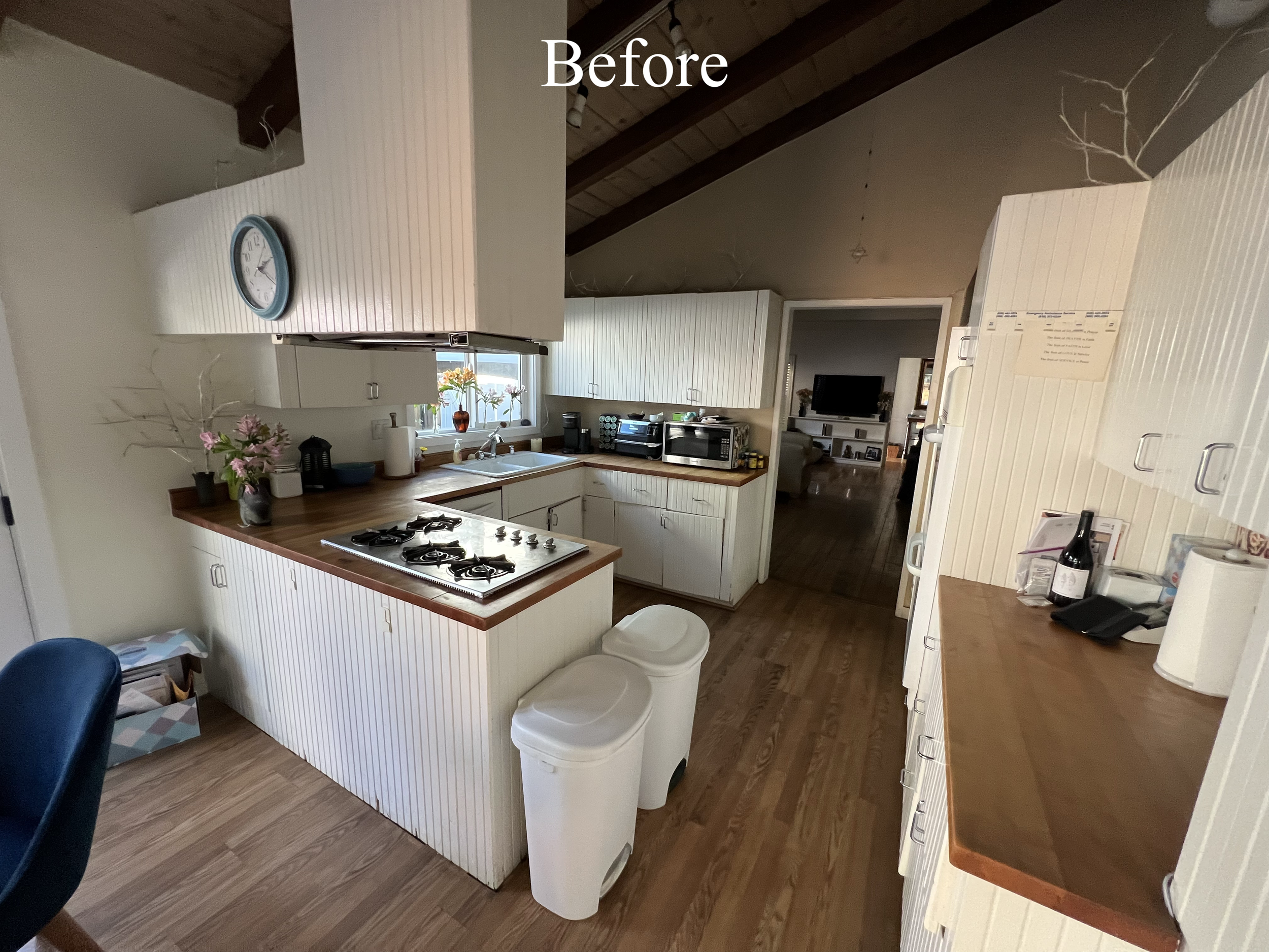 The kitchen has white cabinets with vertical paneling, a wooden countertop, and a gas stove. There is a clock on the wall, a window with potted flowers, and kitchen appliances like a microwave and coffee maker. Two white trash bins are in front of th