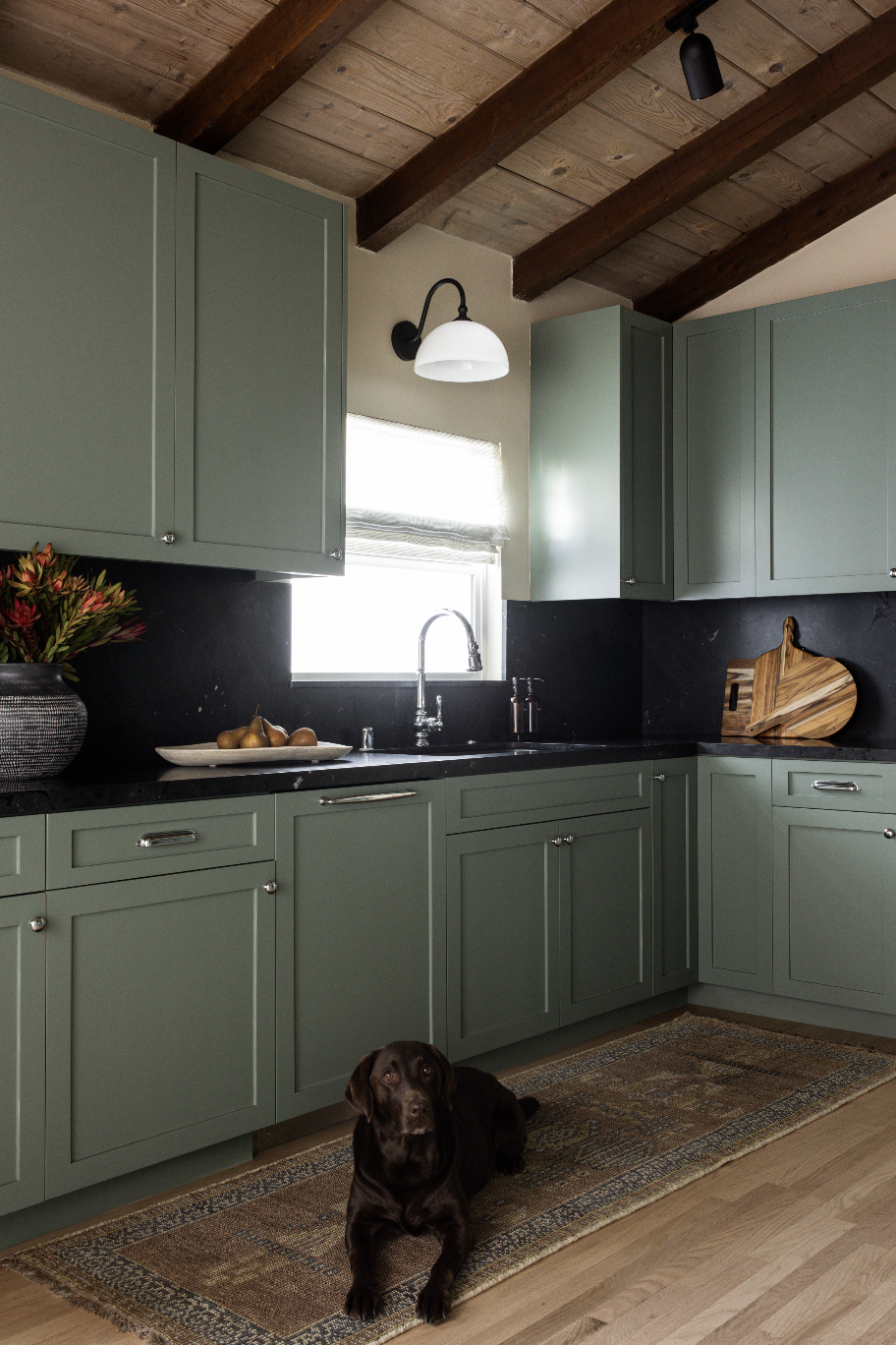 Kitchen with green cabinets, black countertop, window above sink, wooden ceiling beams, ceiling light, cutting board, vase with flowers, dog sitting on rug.