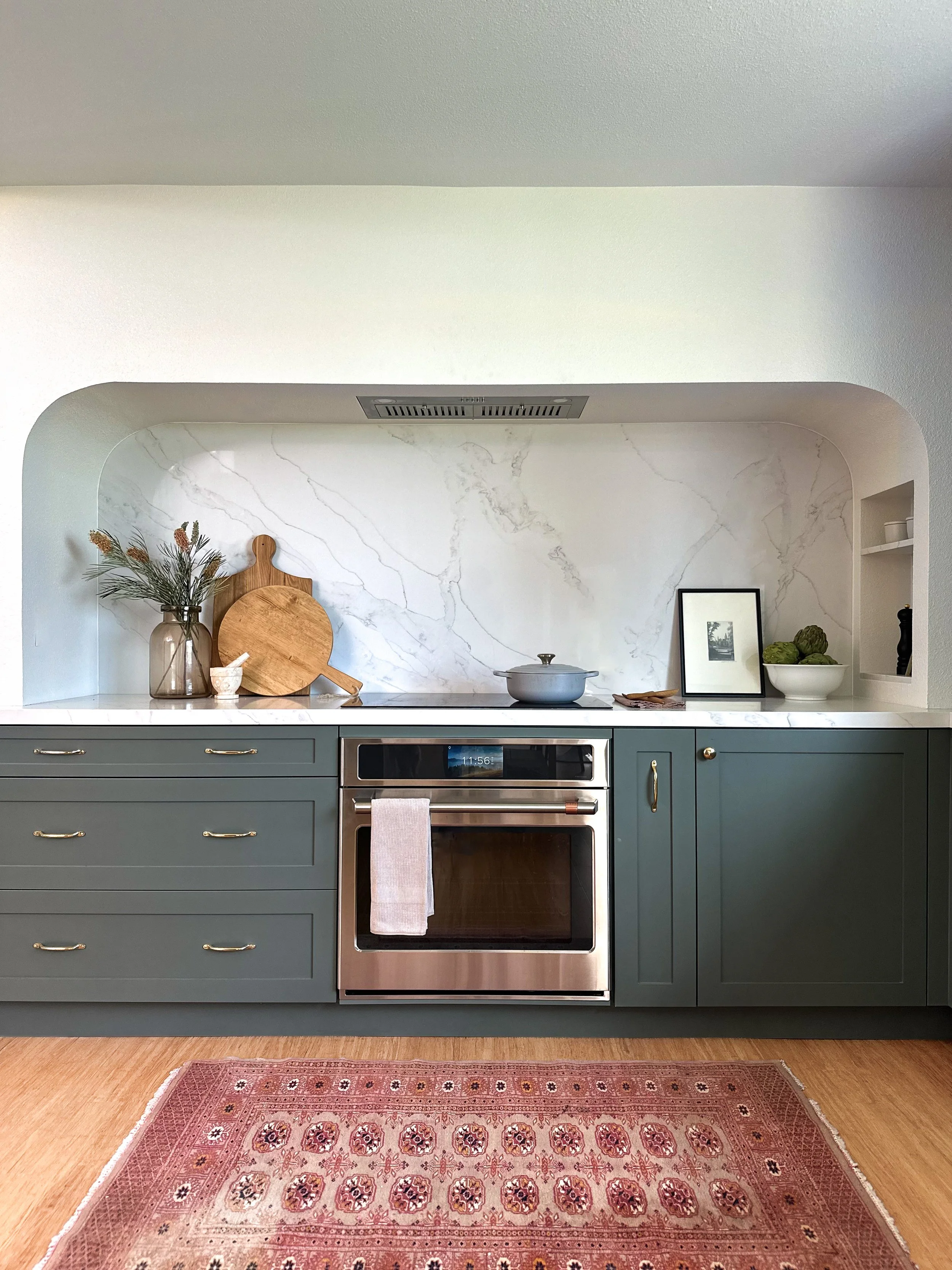 Kitchen with green cabinets, white marble backsplash, and wooden accents. Items on the counter include a glass jar with flowers, a wooden cutting board, a gray pot, a framed picture, and a bowl of green artichokes. A pink patterned area rug is on the