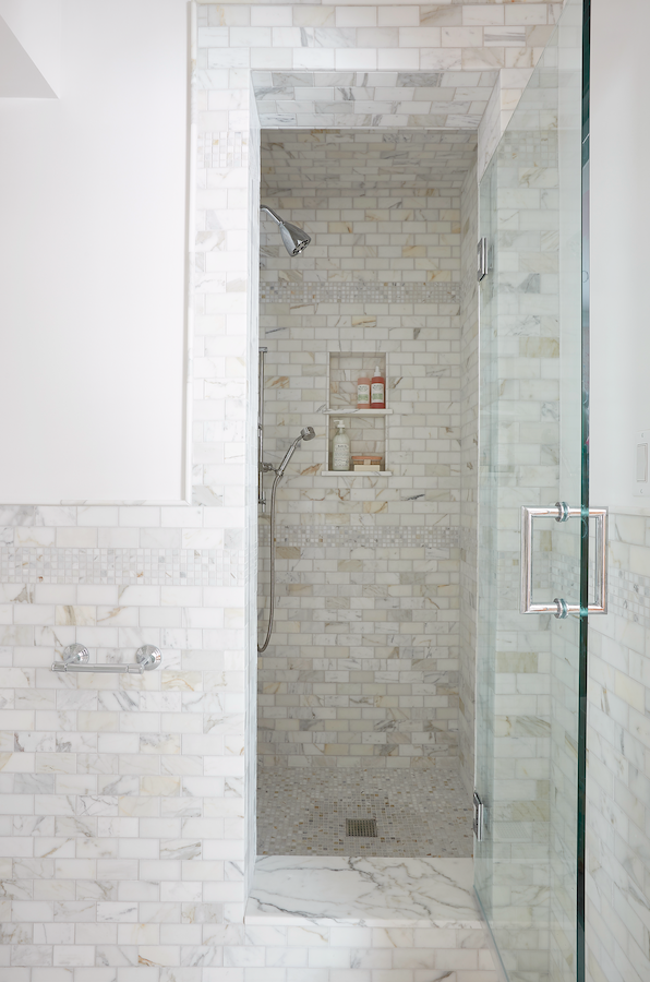 A modern walk-in shower with white marble tiles, a built-in shelf with bottles, a handheld showerhead, and a glass door.