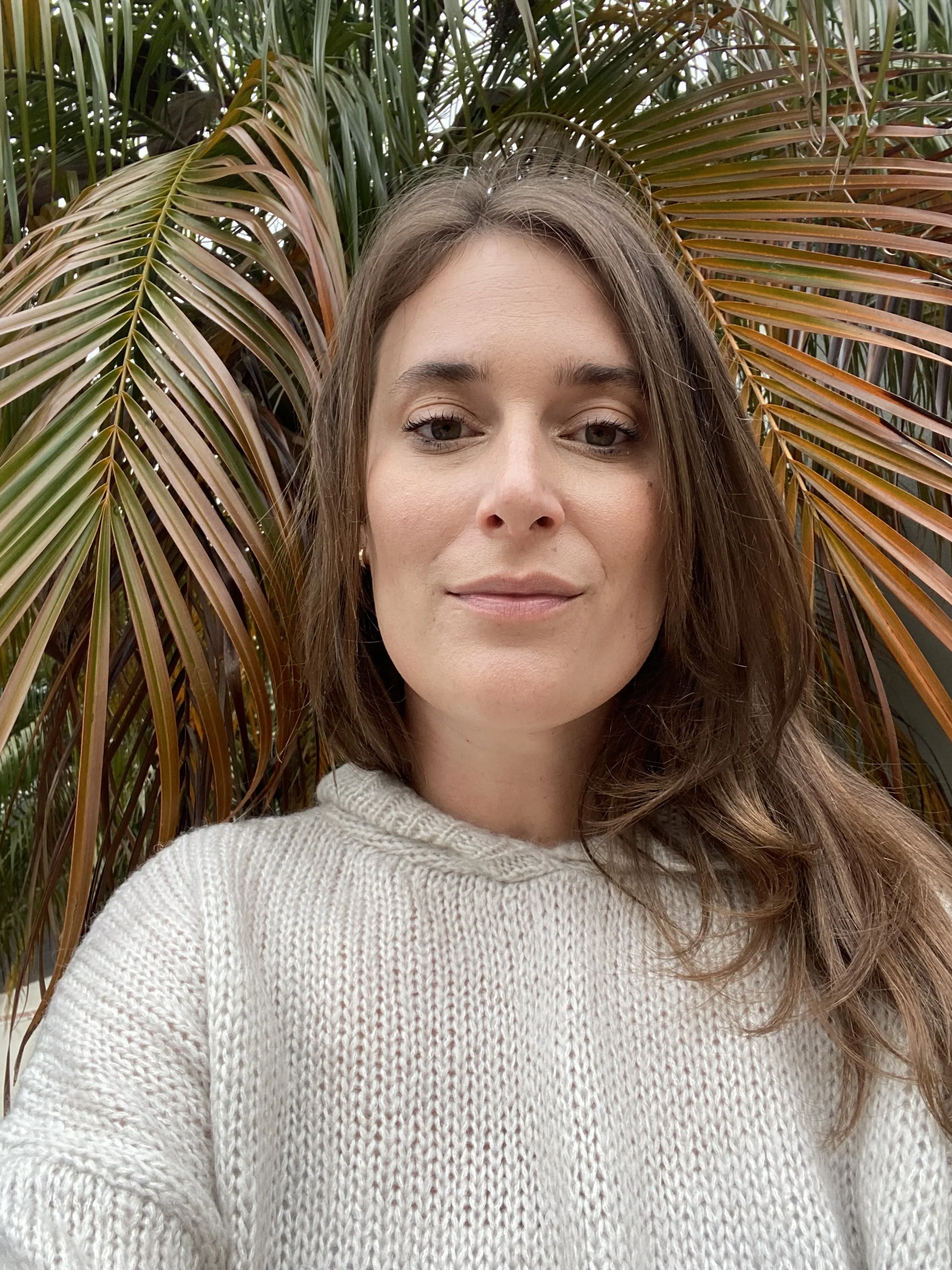 A woman with brown hair and light makeup taking a selfie outdoors in front of brown and green palm leaves, wearing a cream-colored knit sweater.