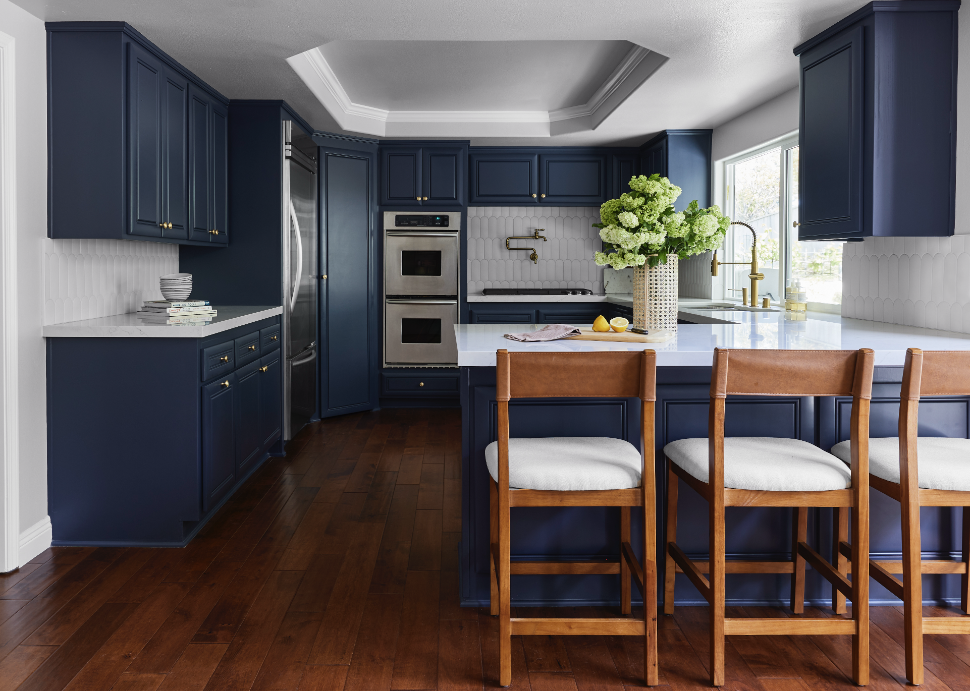 A modern kitchen with navy blue cabinets, white countertops, a white tiled backsplash, a large window with gold fixtures, a large bouquet of white-green flowers in a woven vase, and brown barstools with white cushions.