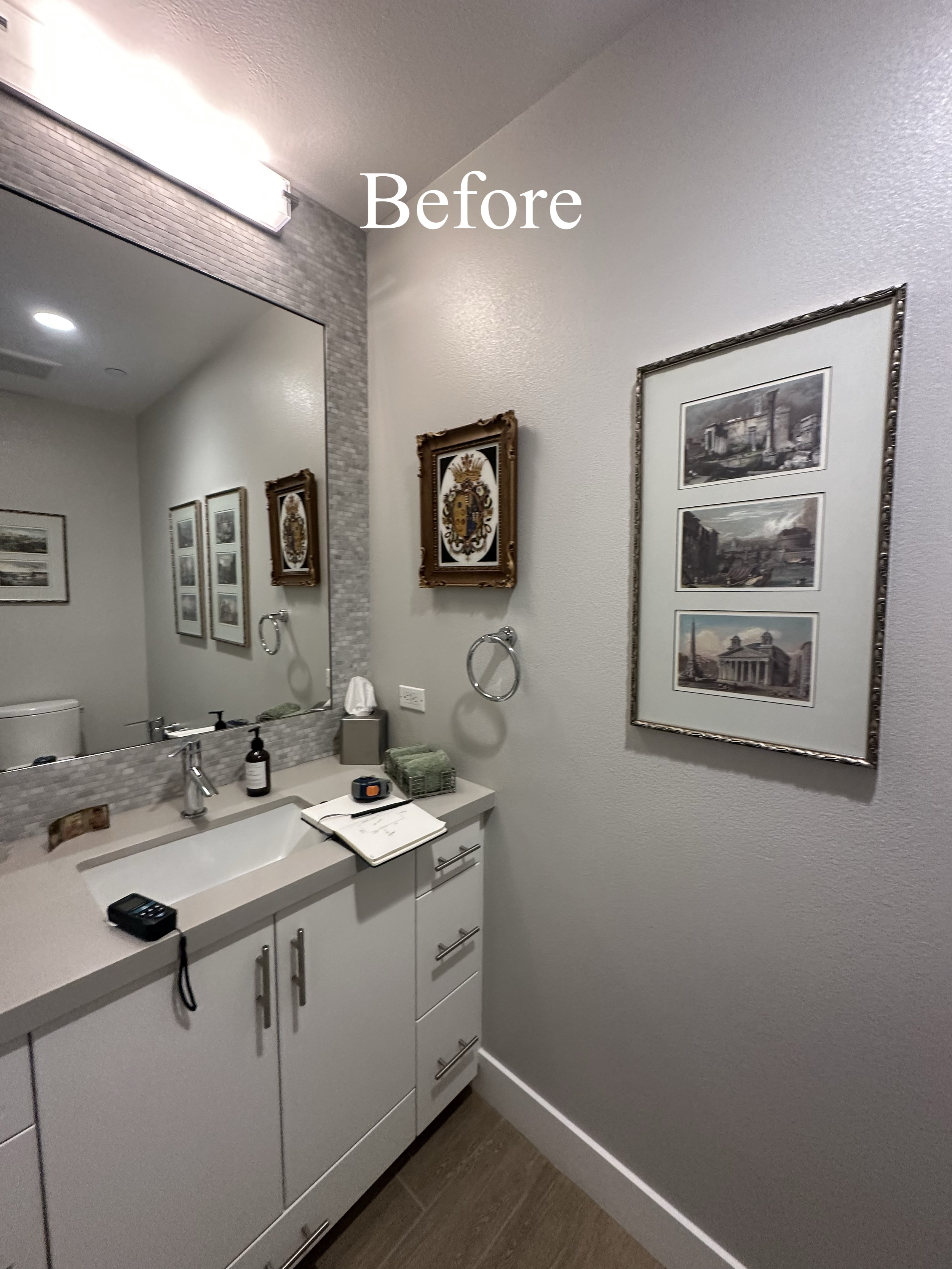 Bathroom with a large mirror, white vanity with drawers, framed artwork on the wall, and a gray textured wall adjacent to a plain wall.