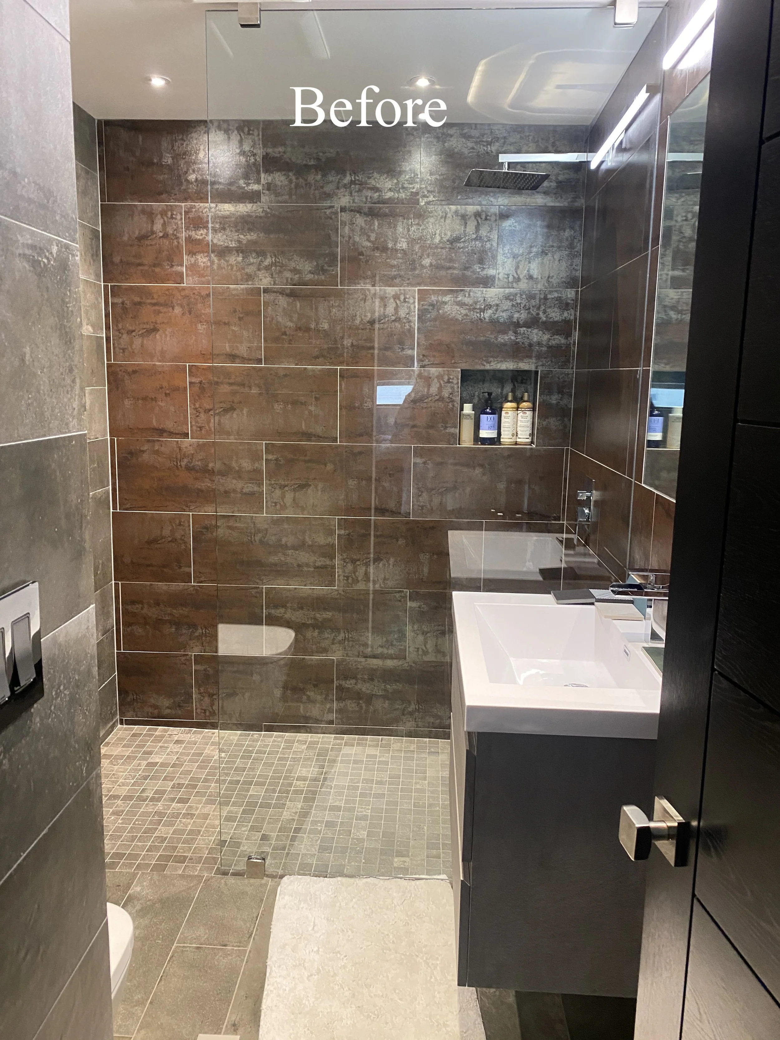 A modern bathroom with a walk-in shower featuring brown tiled walls and a small built-in shelf holding toiletries, a white sink with a black cabinet, and a large mirror.
