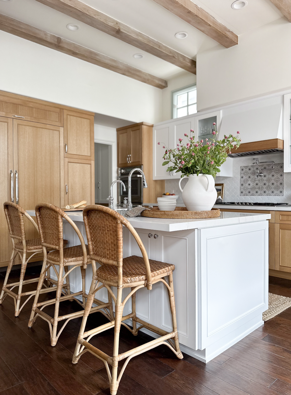 Modern kitchen with white and wood cabinetry, a white island with a vase of pink flowers, wicker bar stools, and stainless steel fixtures.