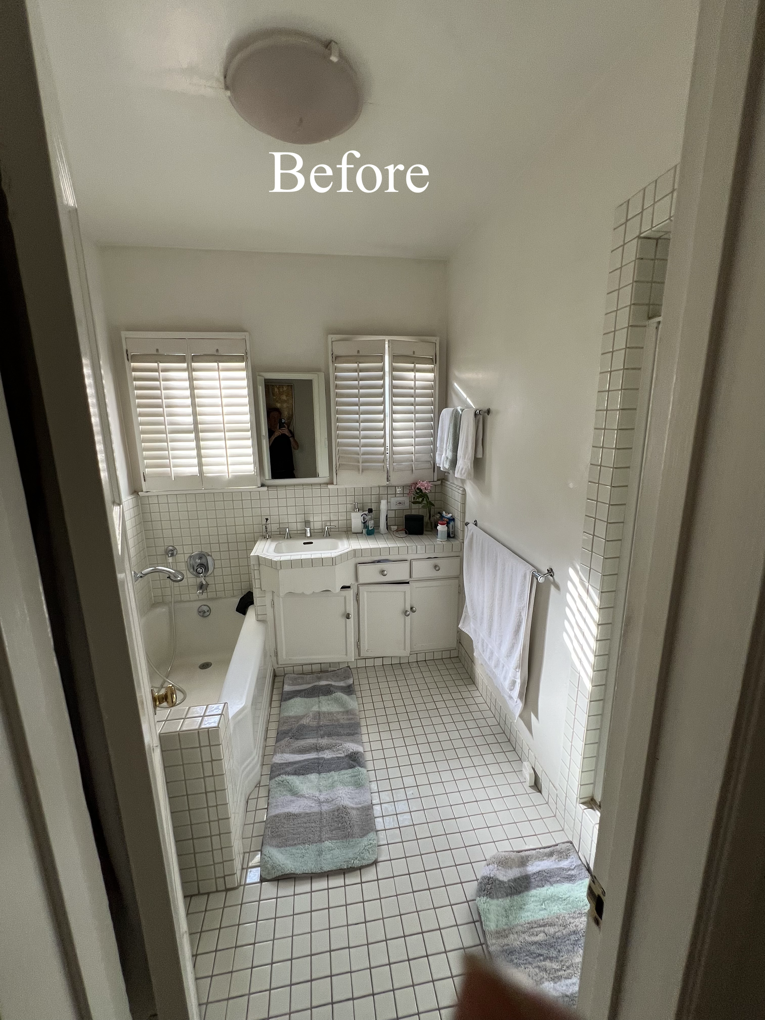 A small vintage bathroom with white tiled floor, a bathtub with a faucet, a vanity with a sink, and a mirror above it. Two small rugs are on the floor. White shutters cover the windows, and there are towels hanging on the wall.