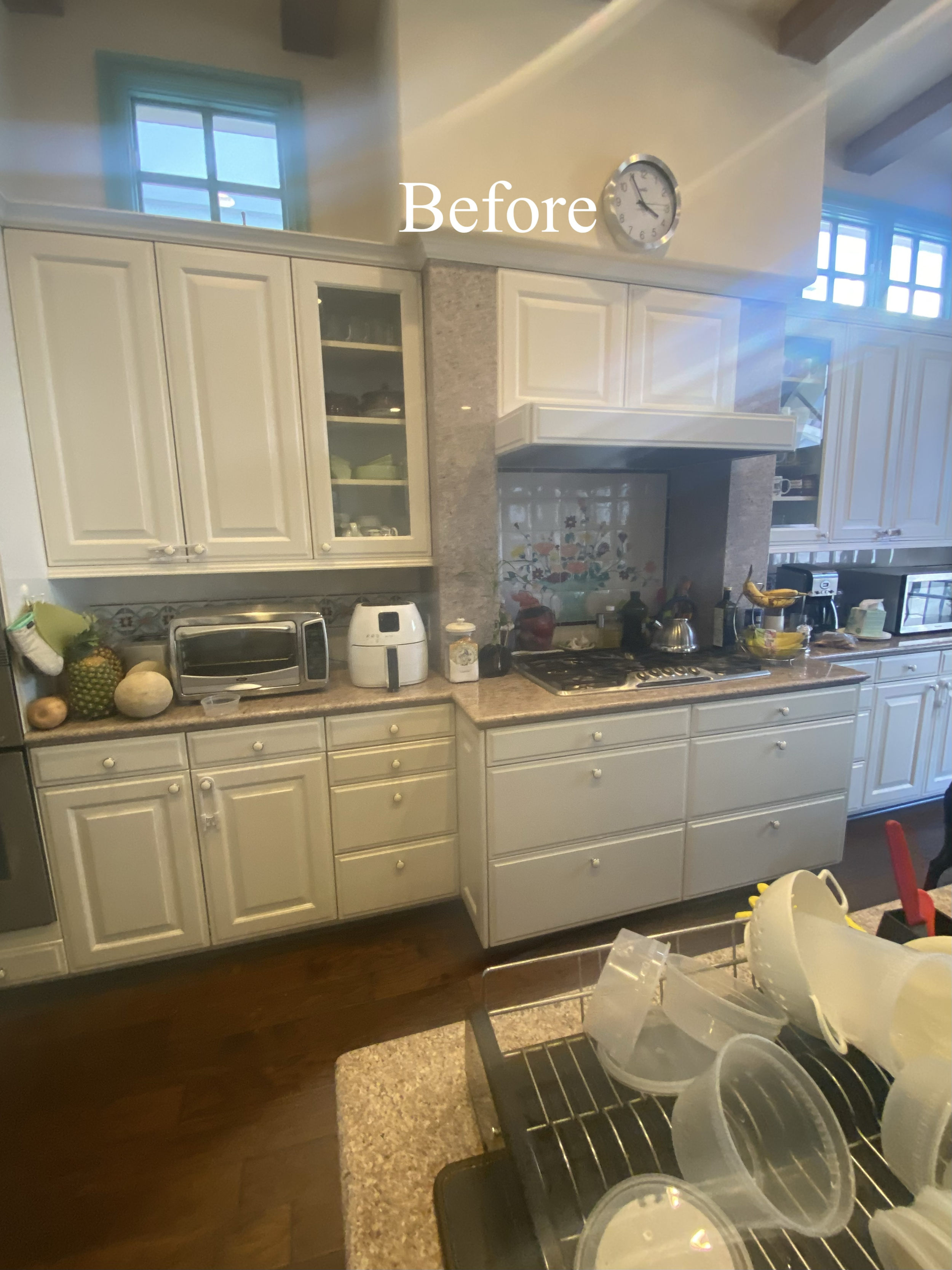 Photo of a kitchen with white cabinets, granite countertops, and various appliances like a microwave, toaster, and air fryer. There are fruits on the counter and a clock on the wall showing 3:10. Cooking utensils and containers are on the stove and i