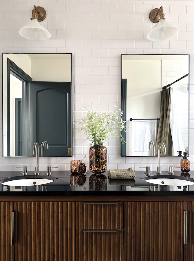 Modern bathroom vanity with double sinks and large mirrors, white brick wall, and decorative items including a vase with flowers, a rolled towel, and soap dispenser. Two wall sconces with white shades above the mirrors, and a window with curtains ref