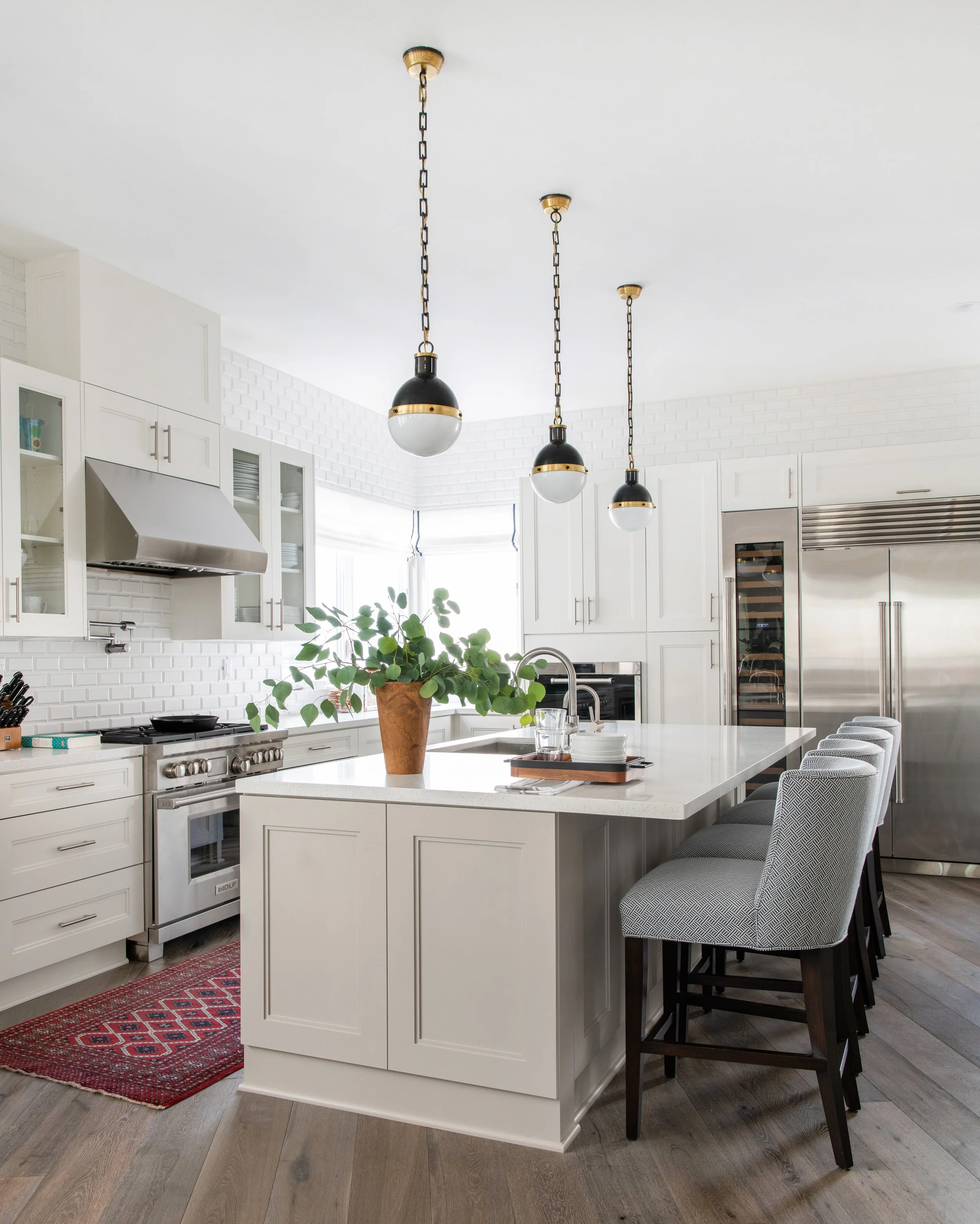 Modern white kitchen with pendant lights, a kitchen island with barstools, stainless steel appliances, and a potted plant on the island.
