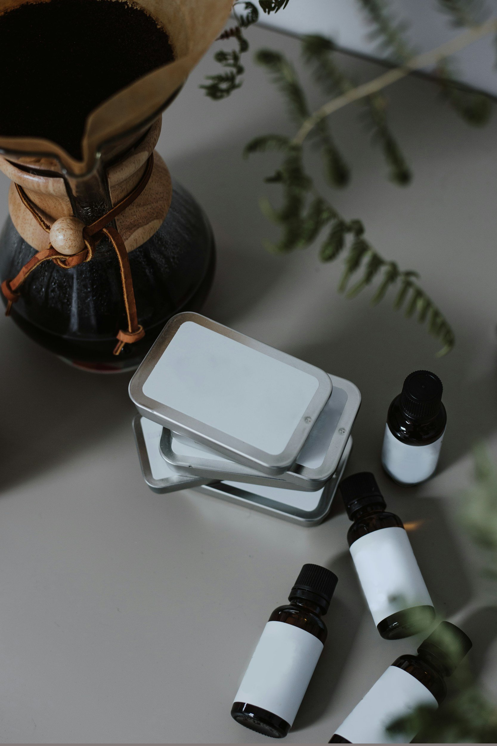 A collection of small brown glass bottles with white labels on a light table, along with a rectangular mirror, a larger bottle with a dropper cap, and a decorative vase with a plant in the background.