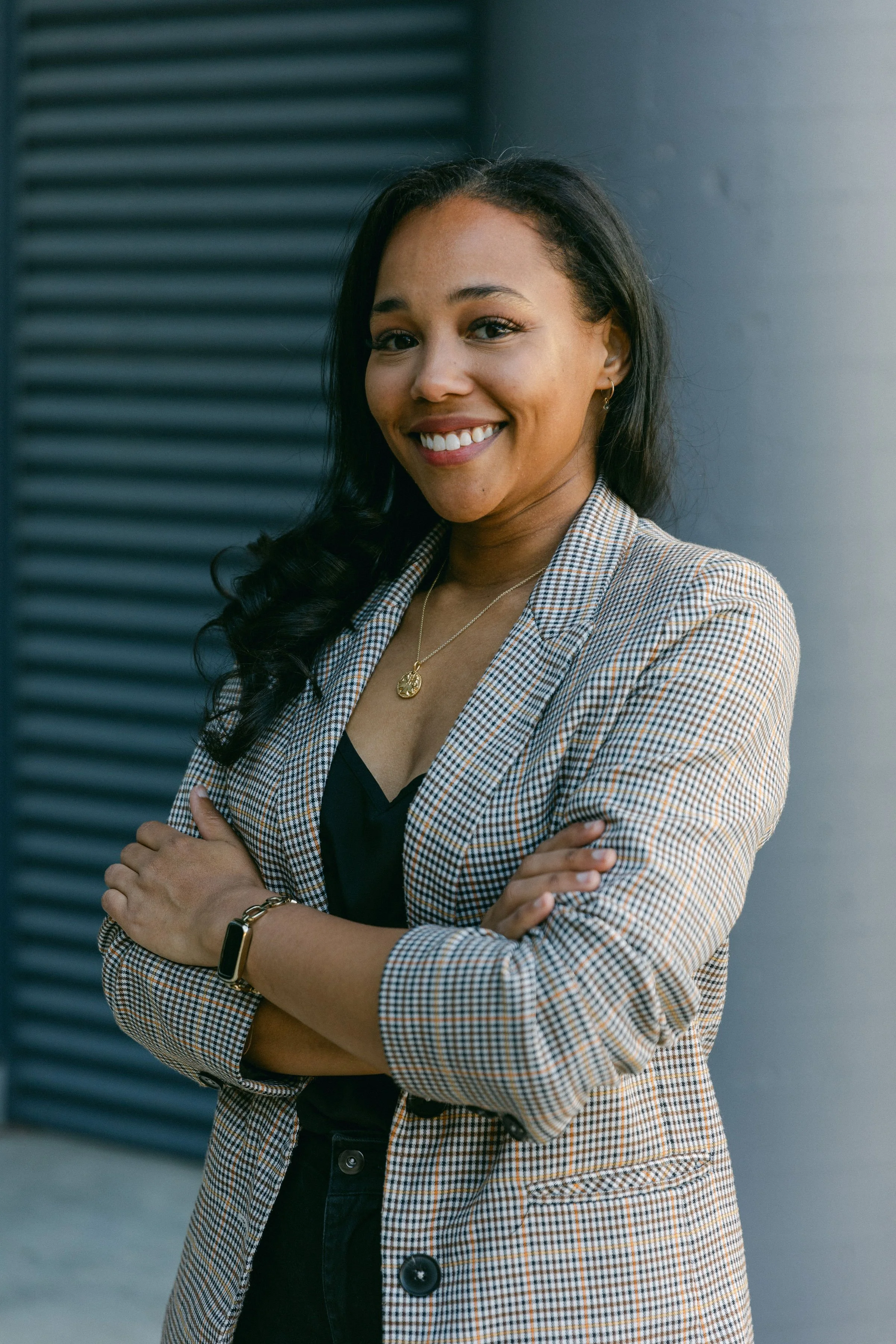 A smiling woman with dark hair in loose curls, wearing a plaid blazer, black top, gold necklace, and a smartwatch, standing outdoors with crossed arms.