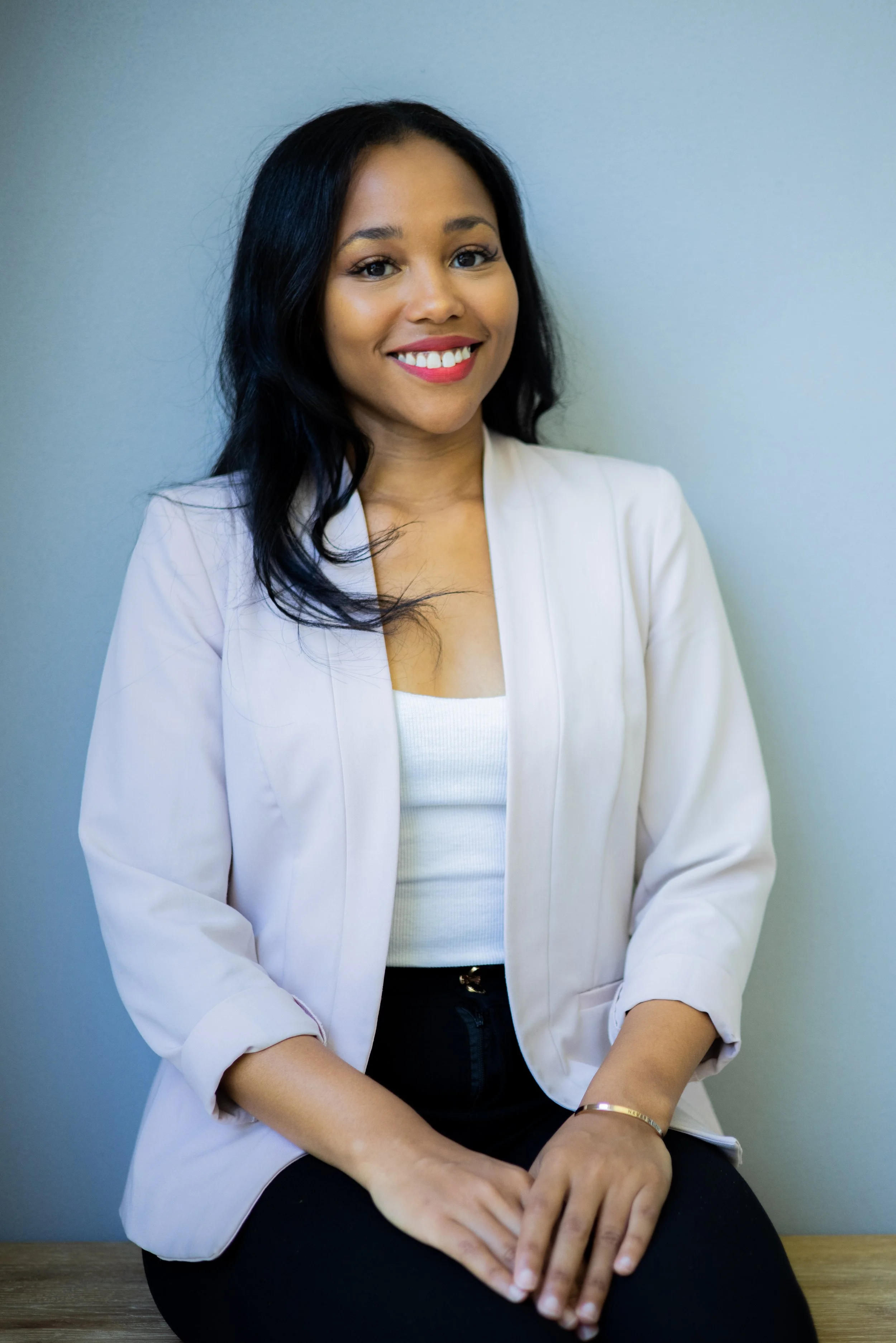 A woman with long black hair, smiling, wearing a white blazer, white top, and black pants, sitting against a light-colored wall.