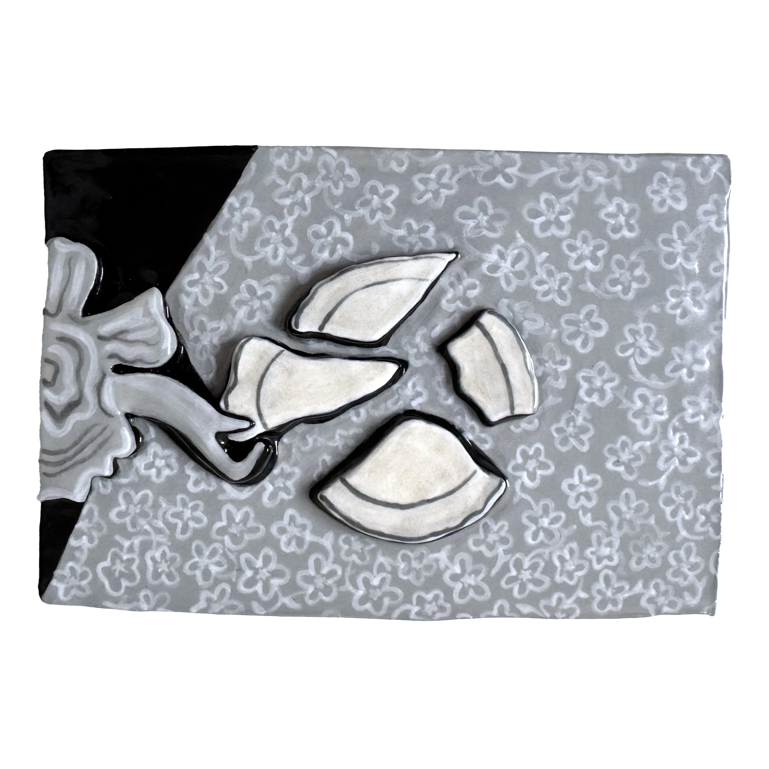 Painted ceramic relief depicting a broken white ceramic plate with black edges on a table covered with a floral-patterned tablecloth.