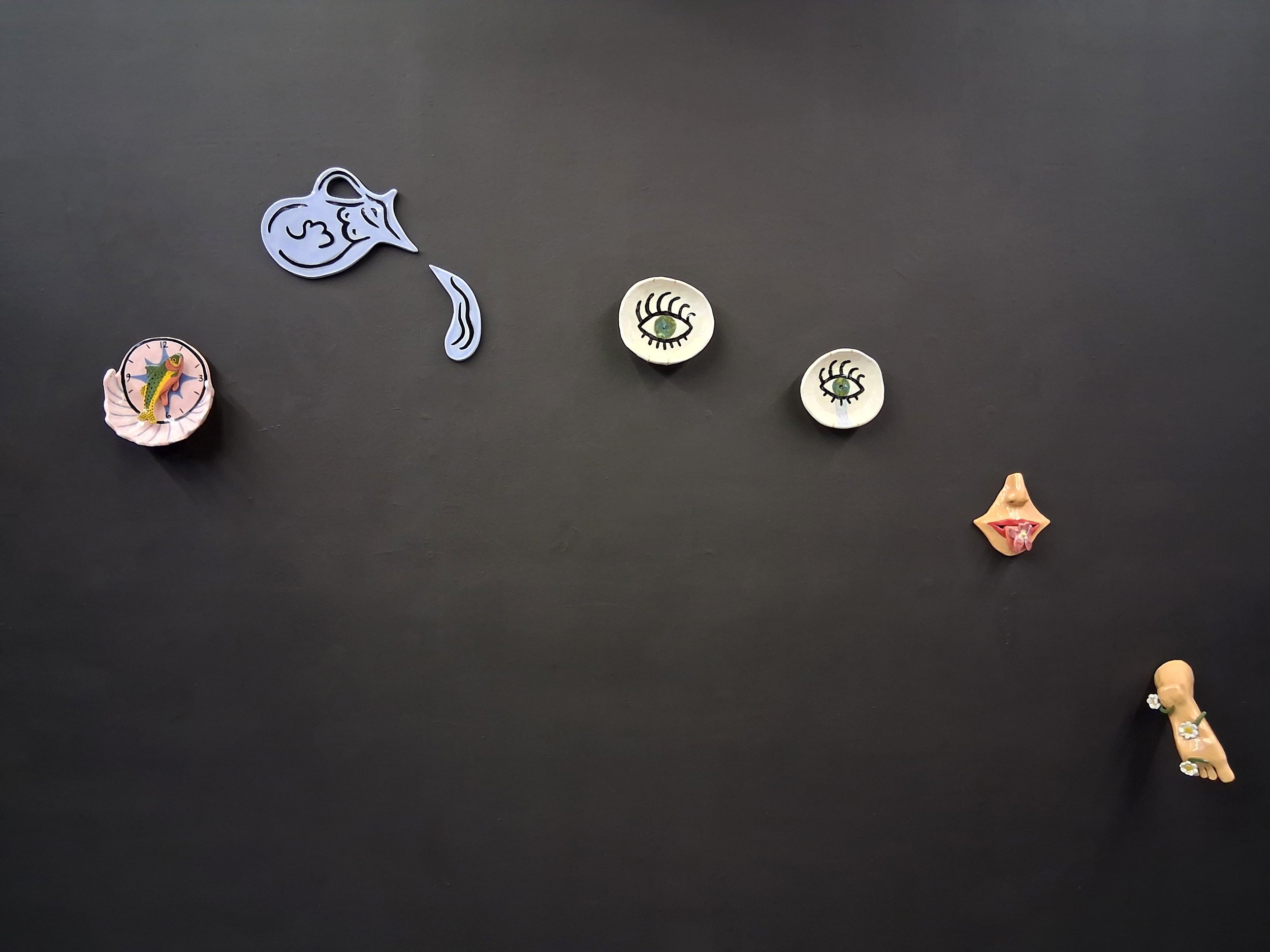 Assorted ceramics hang on a black gallery wall, including a clock with a fish, a water vase, eye-themed bowls, a partial face holding a flower out on its tongue, and a foot covered in daisies.