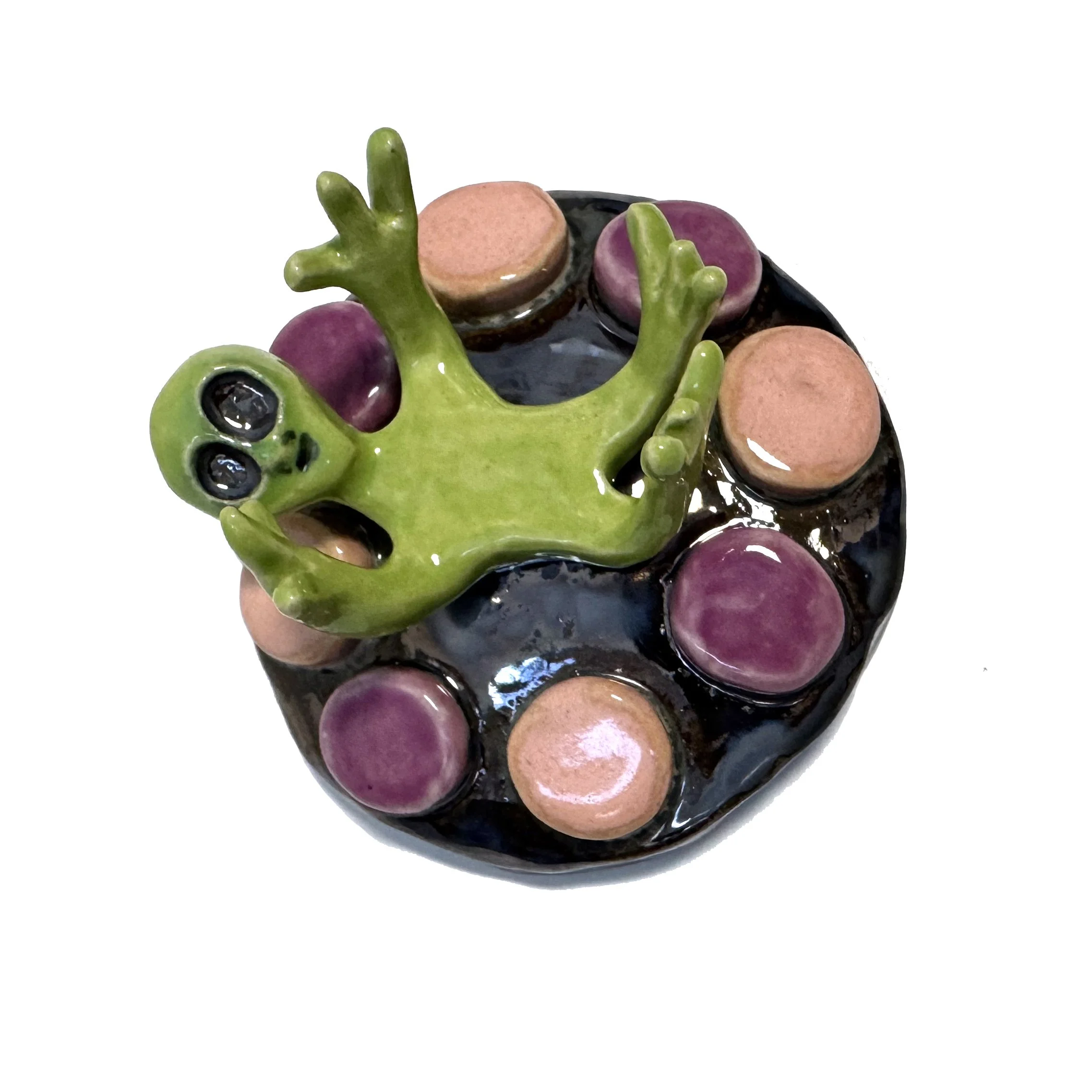 Ceramic figurine of a green alien on a reflective spaceship with pink and purple circular accents.