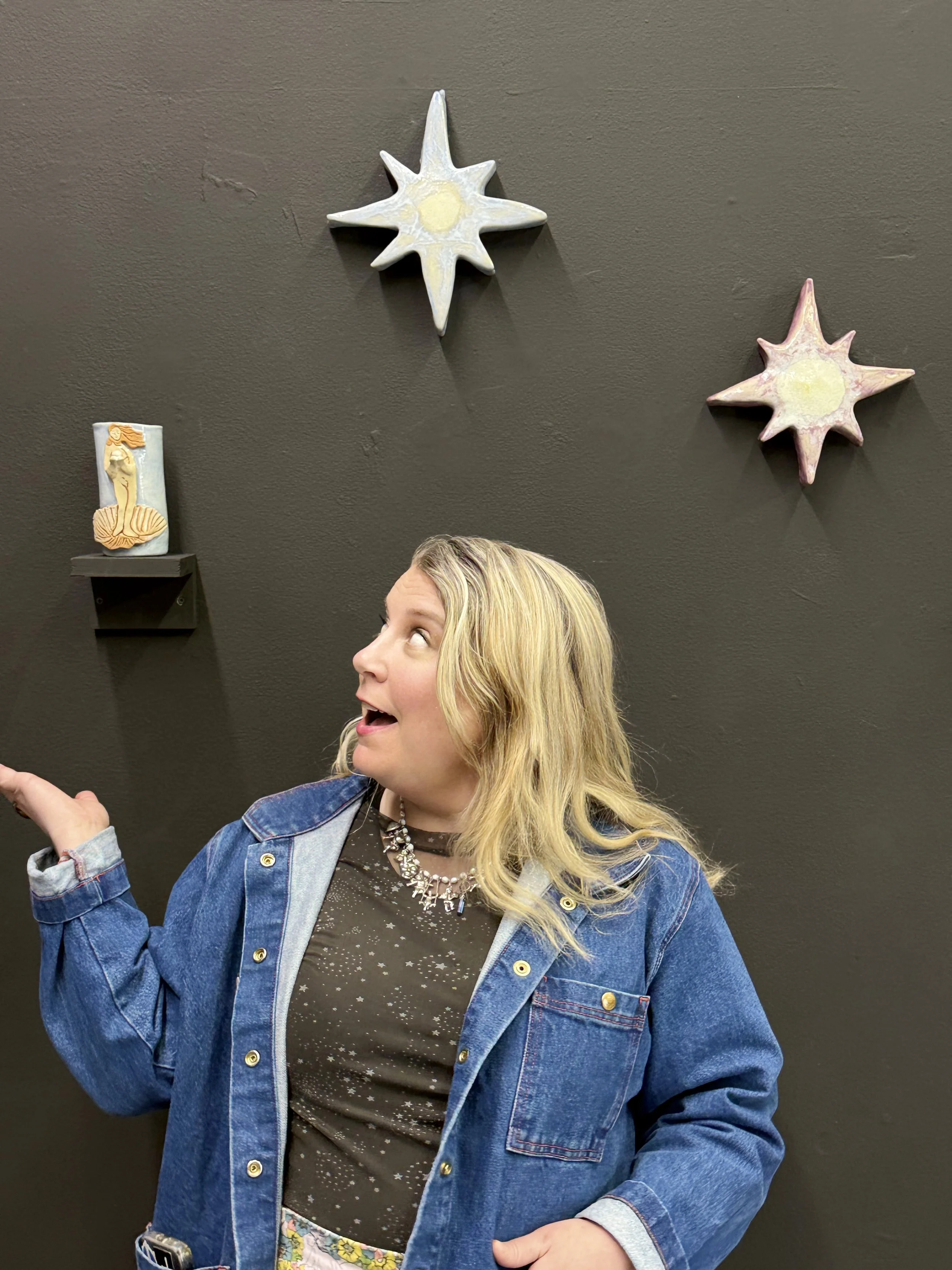 A woman with long blond hair wearing a jean jacket, a shirt with constellations, smiles up at her ceramic Venus cup resting on a shelf on a black wall. Two ceramic stars float above her head.