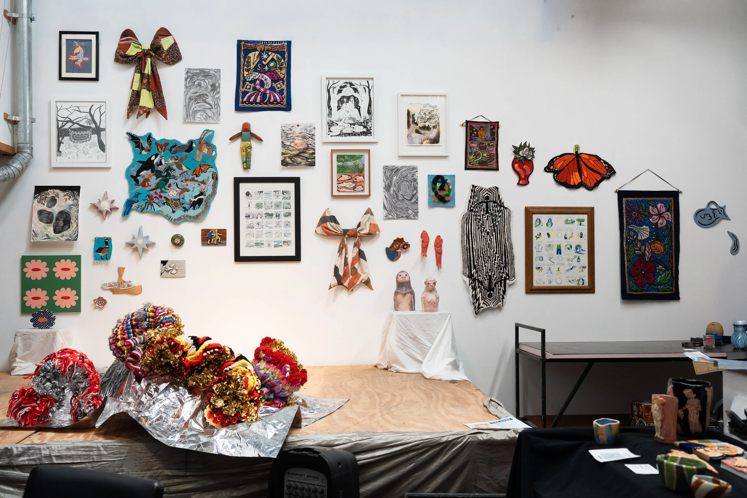 An art gallery wall displaying various framed paintings, textile artworks, and sculptures, with some colorful, abstract, and nature-inspired pieces; foreground shows an installation of multicolored crochet sculptures on a stage.