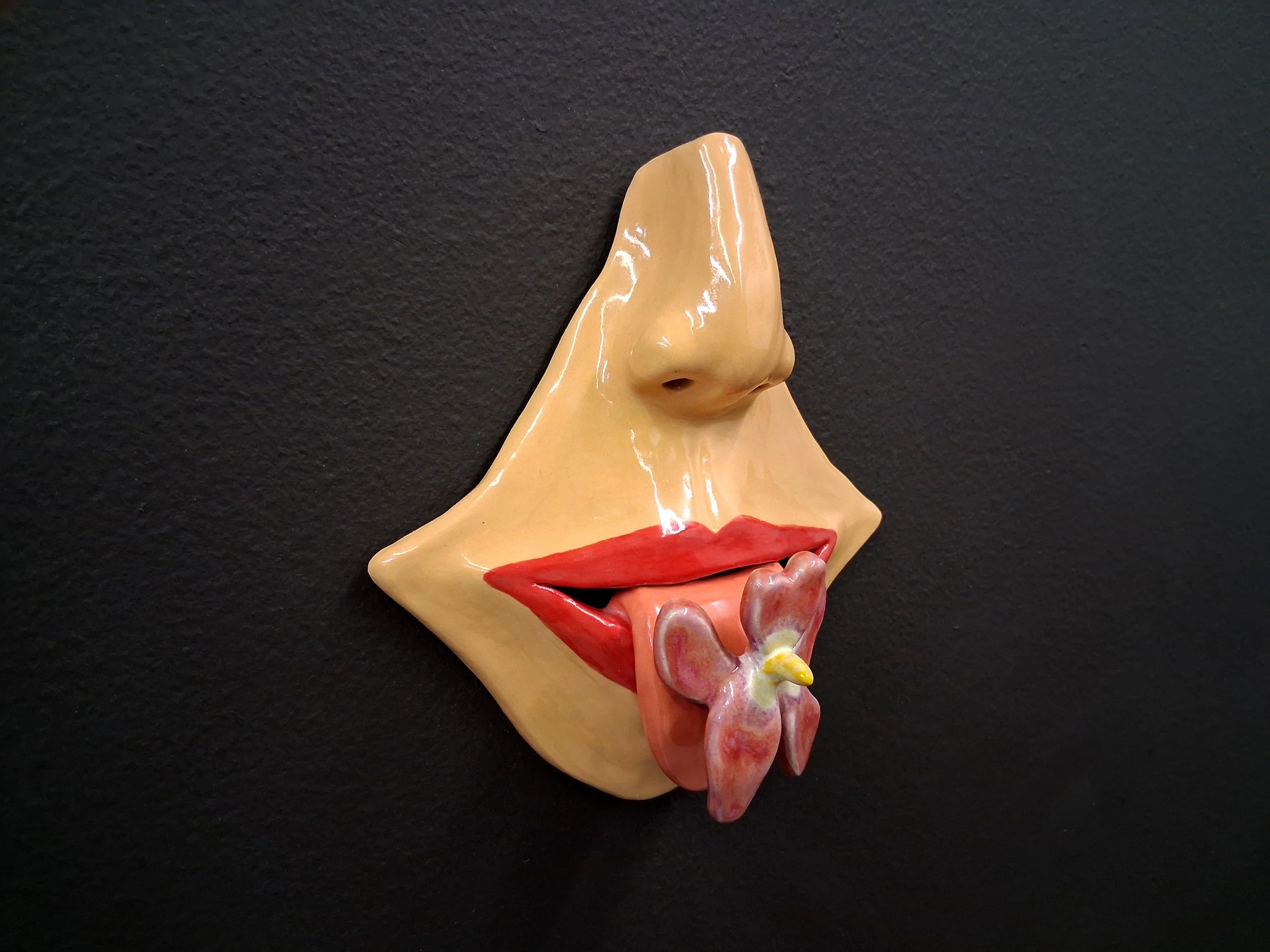 A surreal sculpture of a human face combining a nose and lips with a flower at the mouth, painted in flesh tones with red lips and black background.