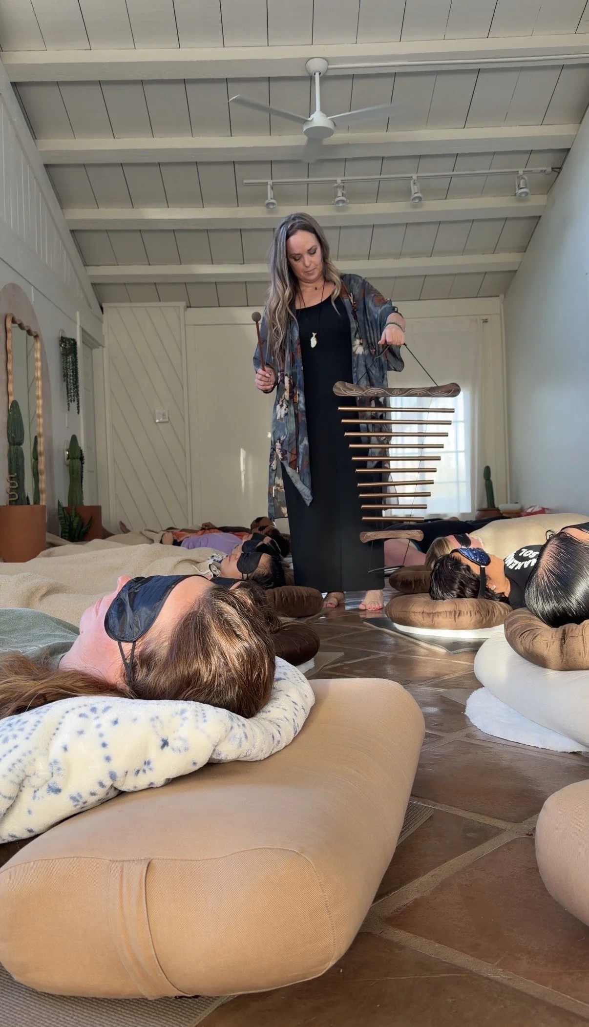 A woman standing among people lying on cushions on the floor, playing a musical instrument in a room with high ceilings, wood beams, and a ceiling fan.