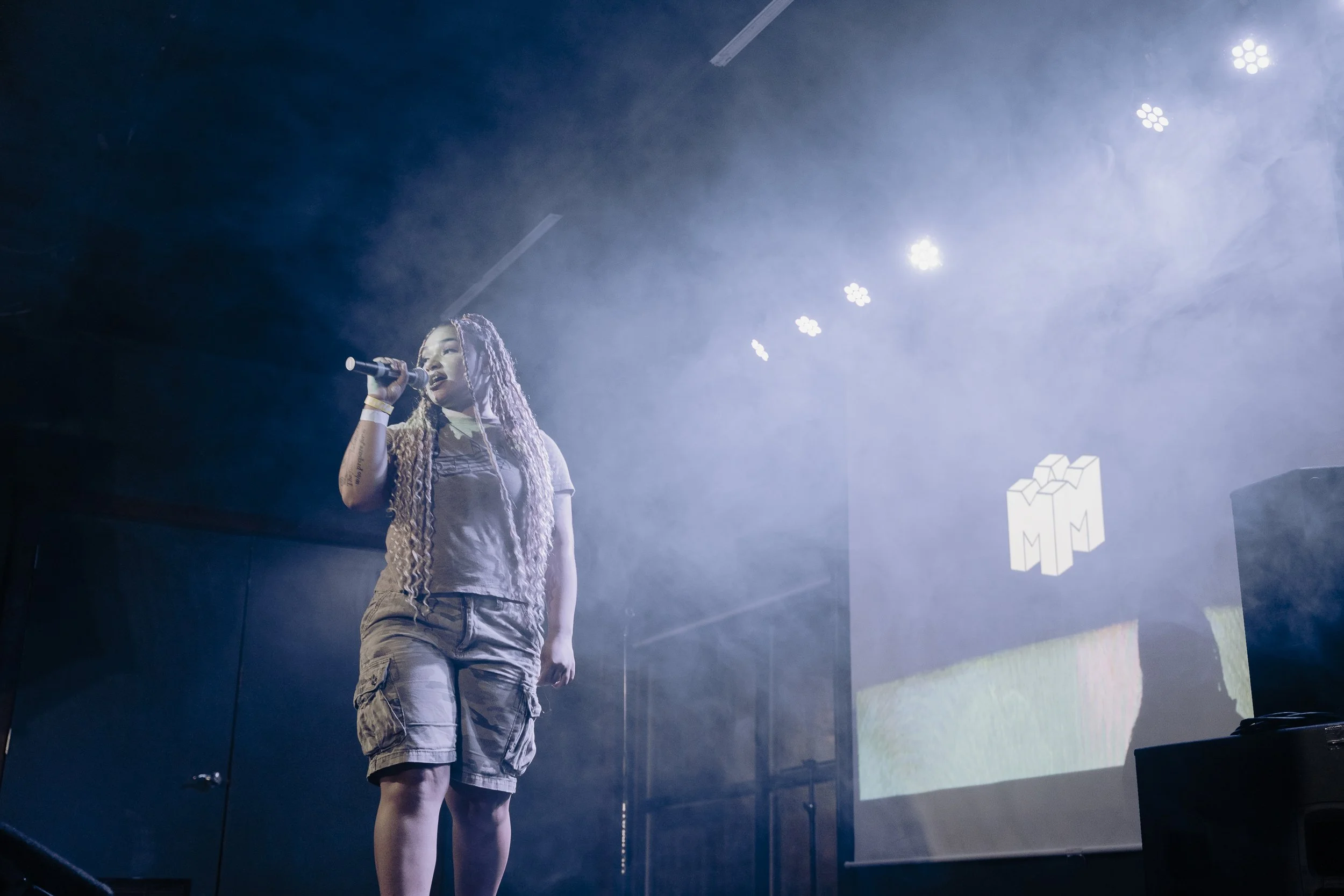 A young woman with long braided hair singing into a microphone on stage with blue lighting and a projection screen in the background.
