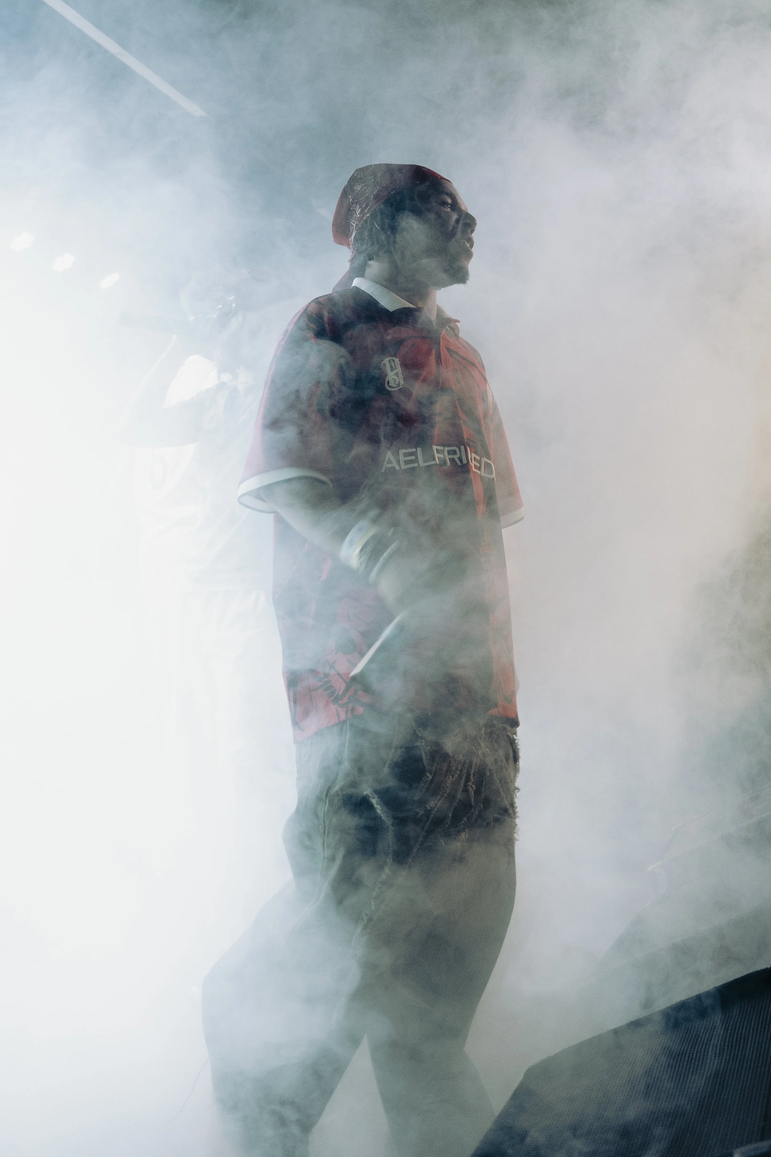 A man standing amidst thick smoke, wearing a red shirt, a person in the background, and the smoke partially obscures the scene.