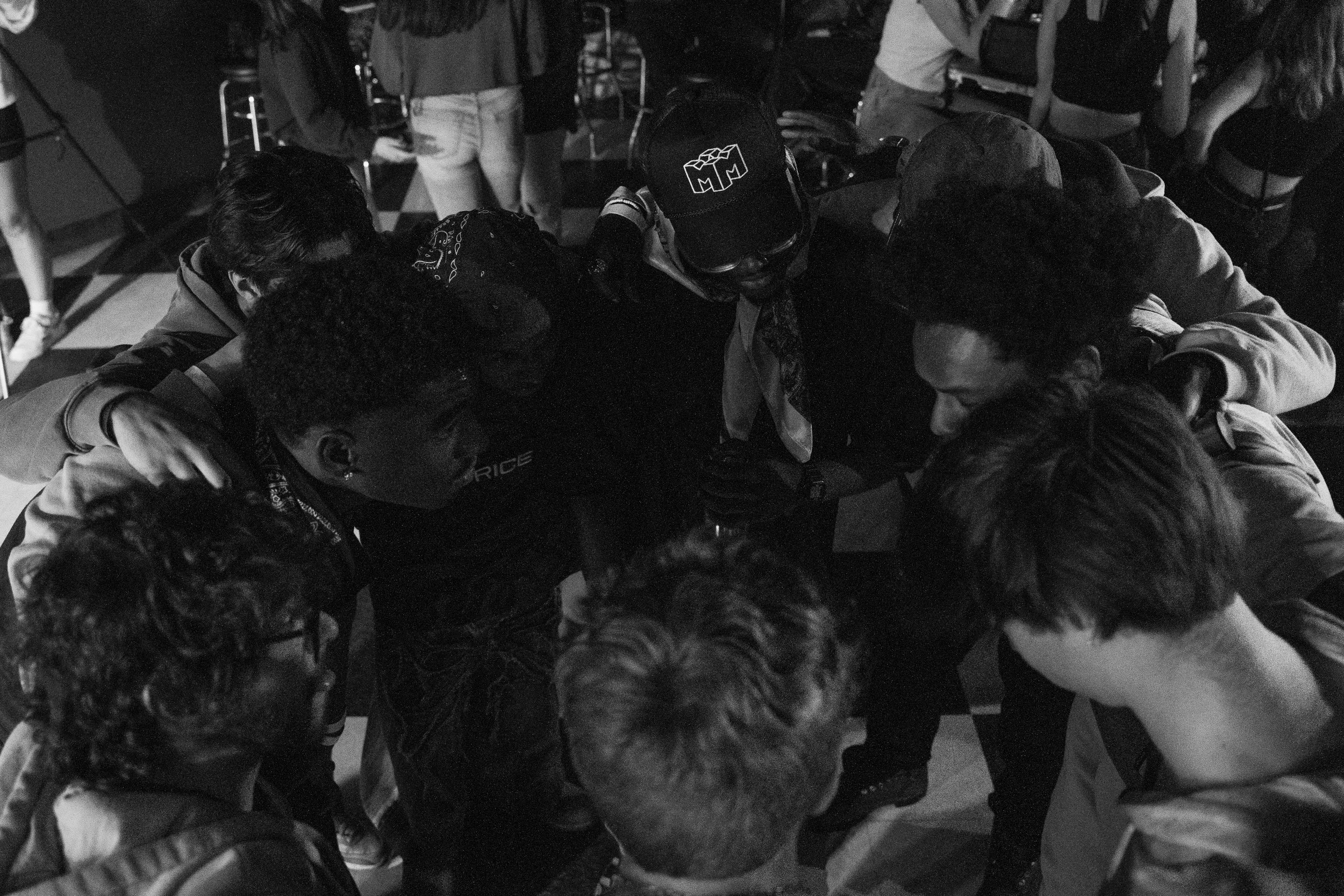 A black and white photo of a group of young people huddled together in a circle, with their arms around each other, in an indoor setting.