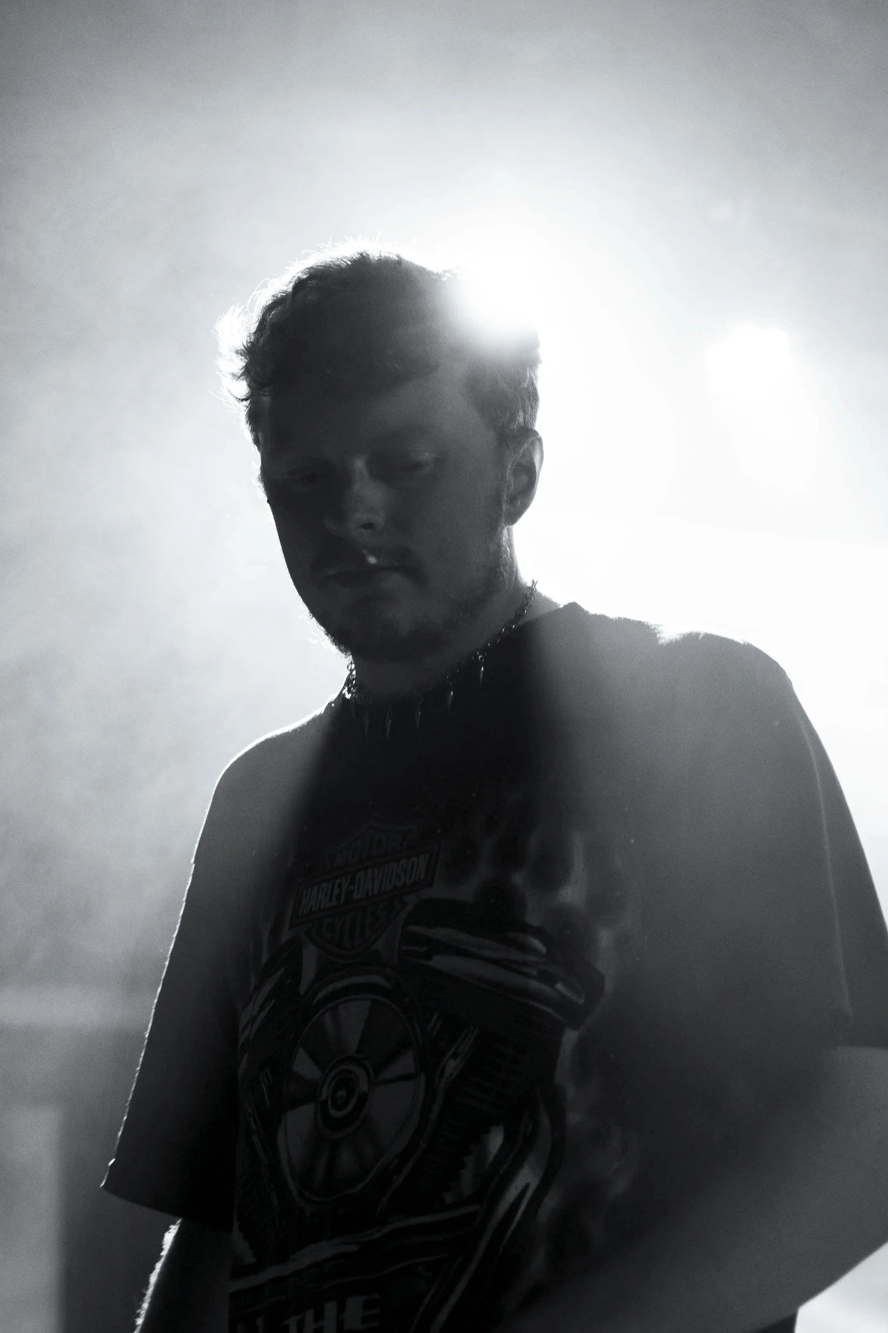 A young man with short hair and facial hair, wearing a black Harley-Davidson shirt and a spiked necklace, stands in a dimly lit environment with bright light behind him, creating a silhouette effect.