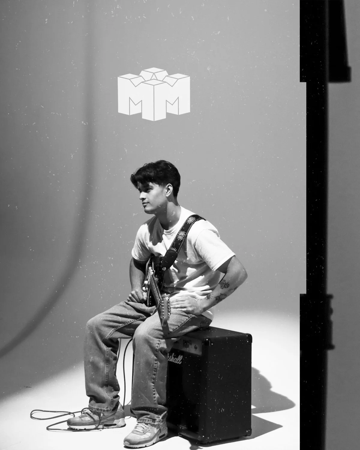 A young person sitting on a stool with a guitar strap over his shoulder, posing against a plain background.