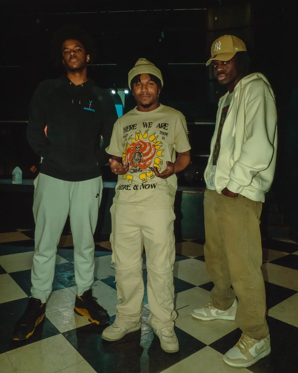 Three young men standing together indoors, dressed in casual streetwear, with a dark background and checkered floor.