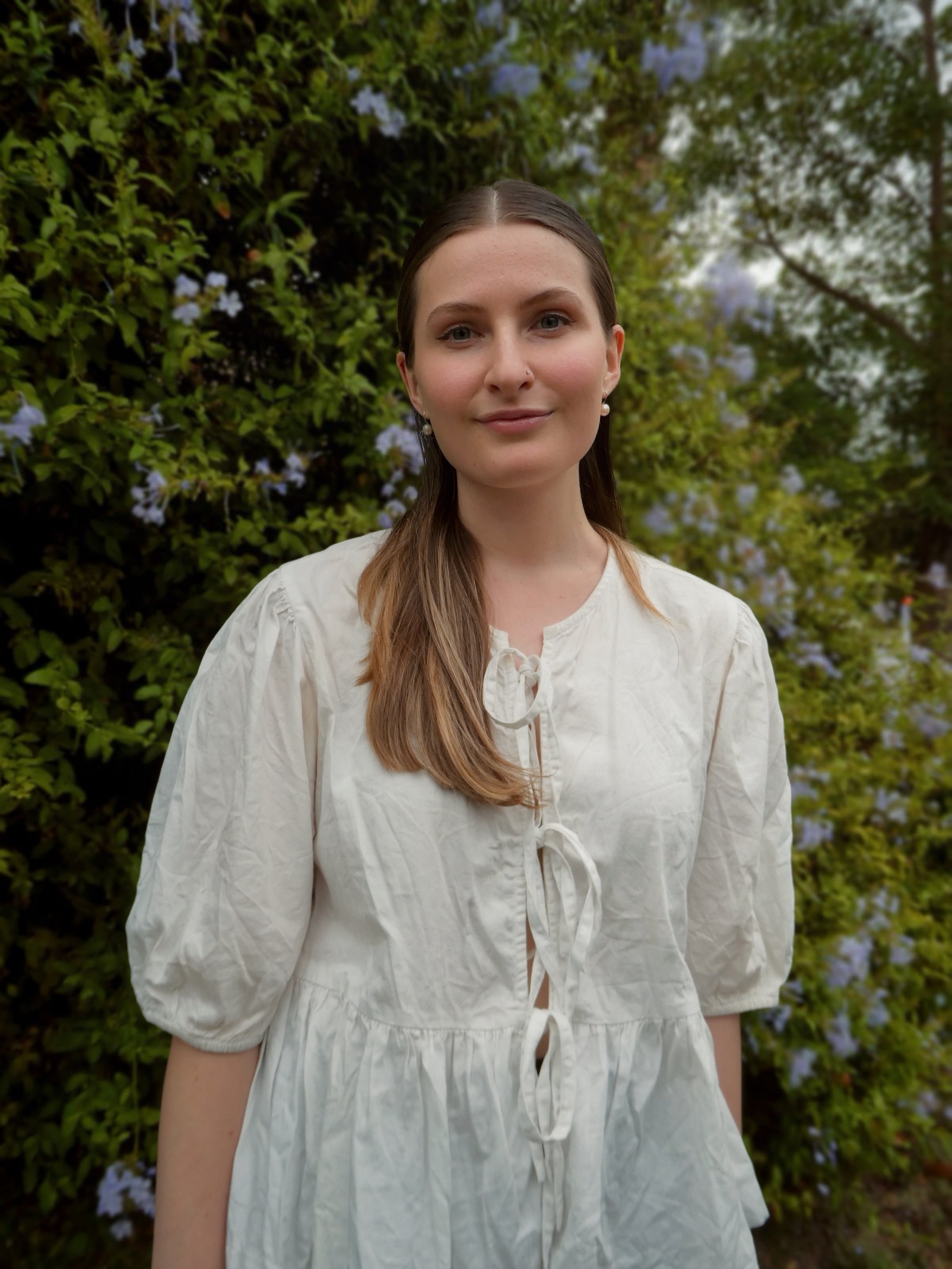 Orana smiles at the camera. She has long brown hair, blue eyes, and a white shirt. She is standing in a garden.
