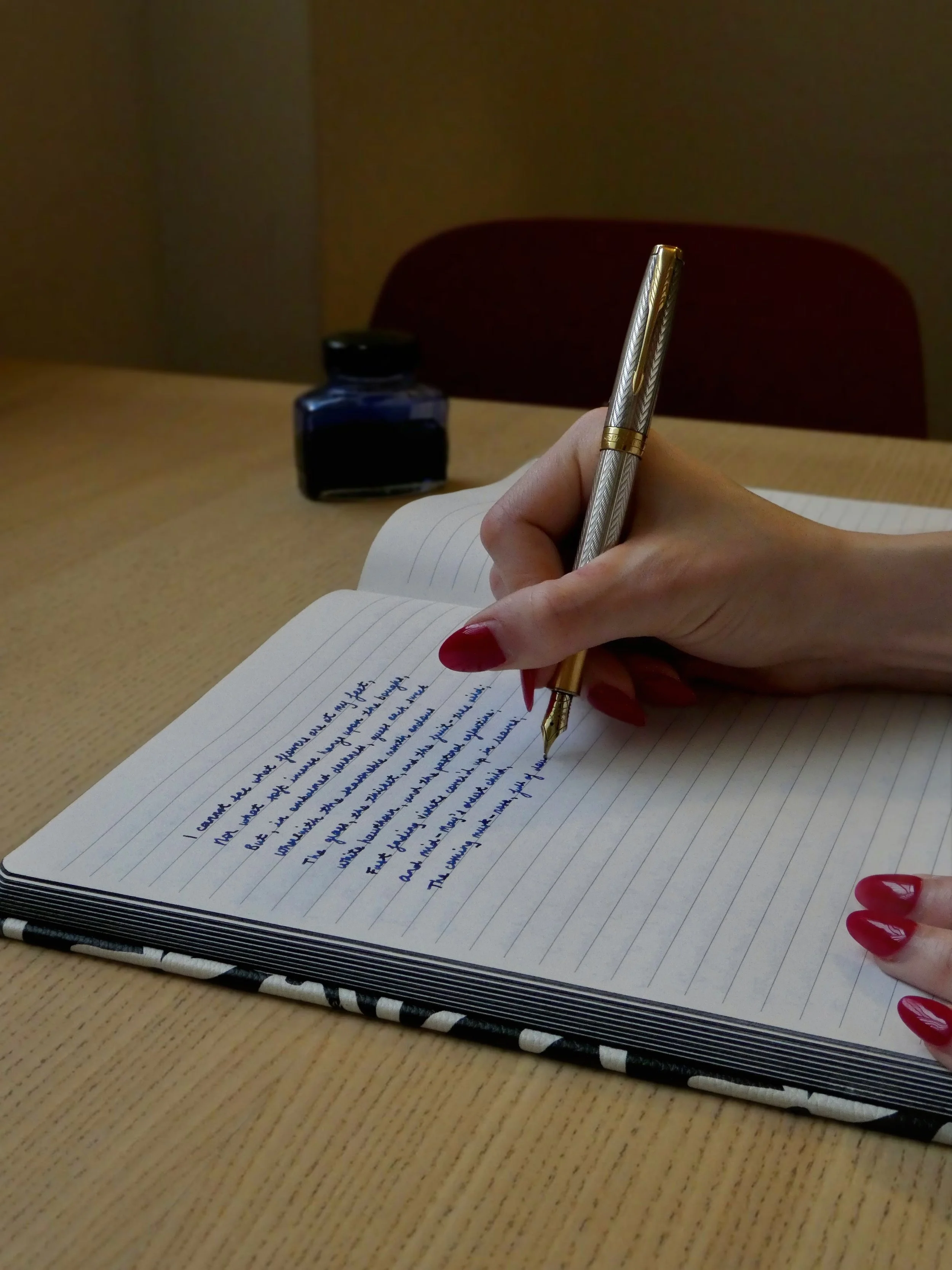 Close up of woman's hand with long red fingernails writing with a silver fountain pen. A blue ink pot is in the background.