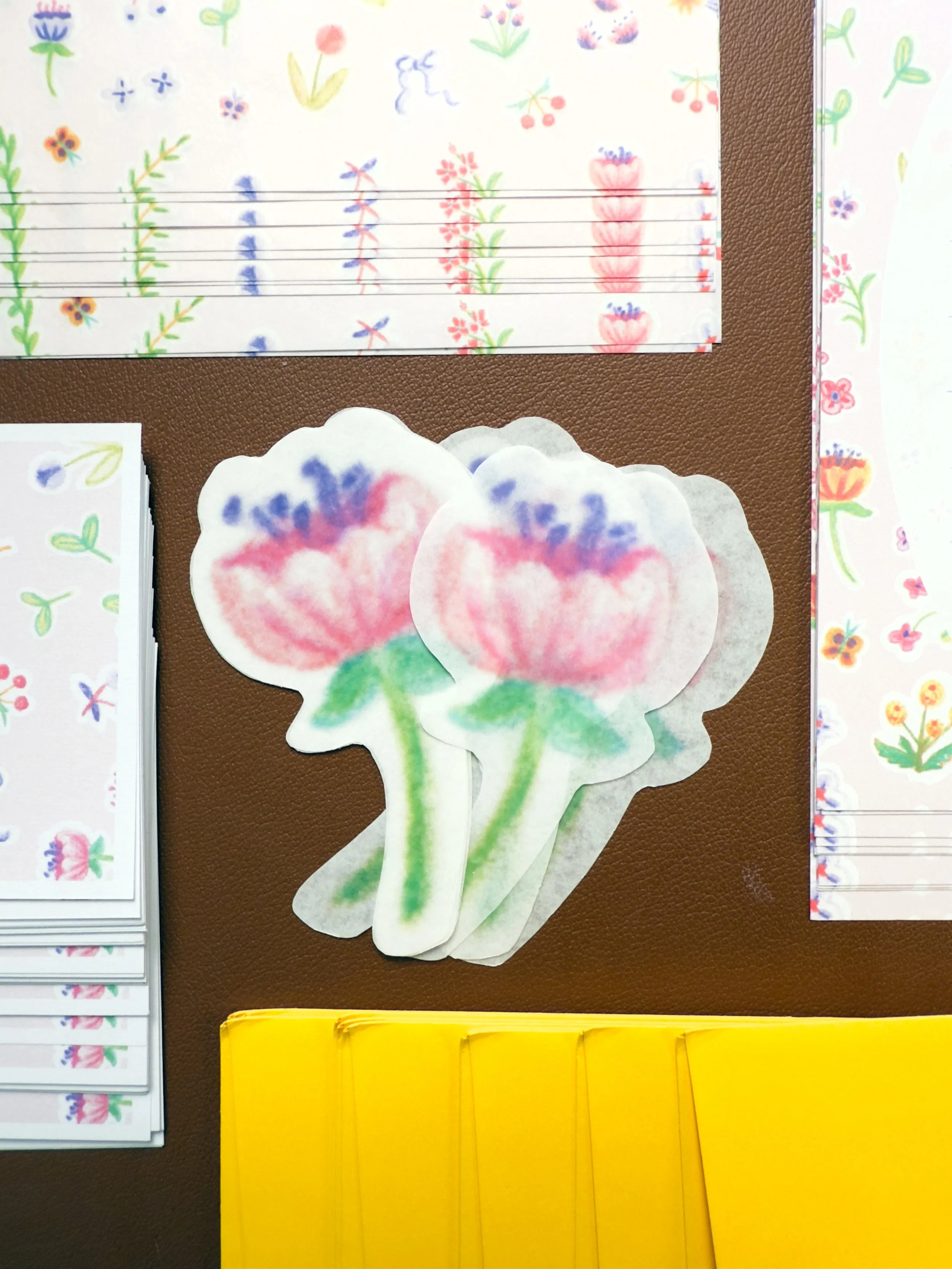 Colorful floral drawings and cutouts on paper, including pink and purple flowers with green stems, and stationery with floral patterns, all arranged on a brown surface.