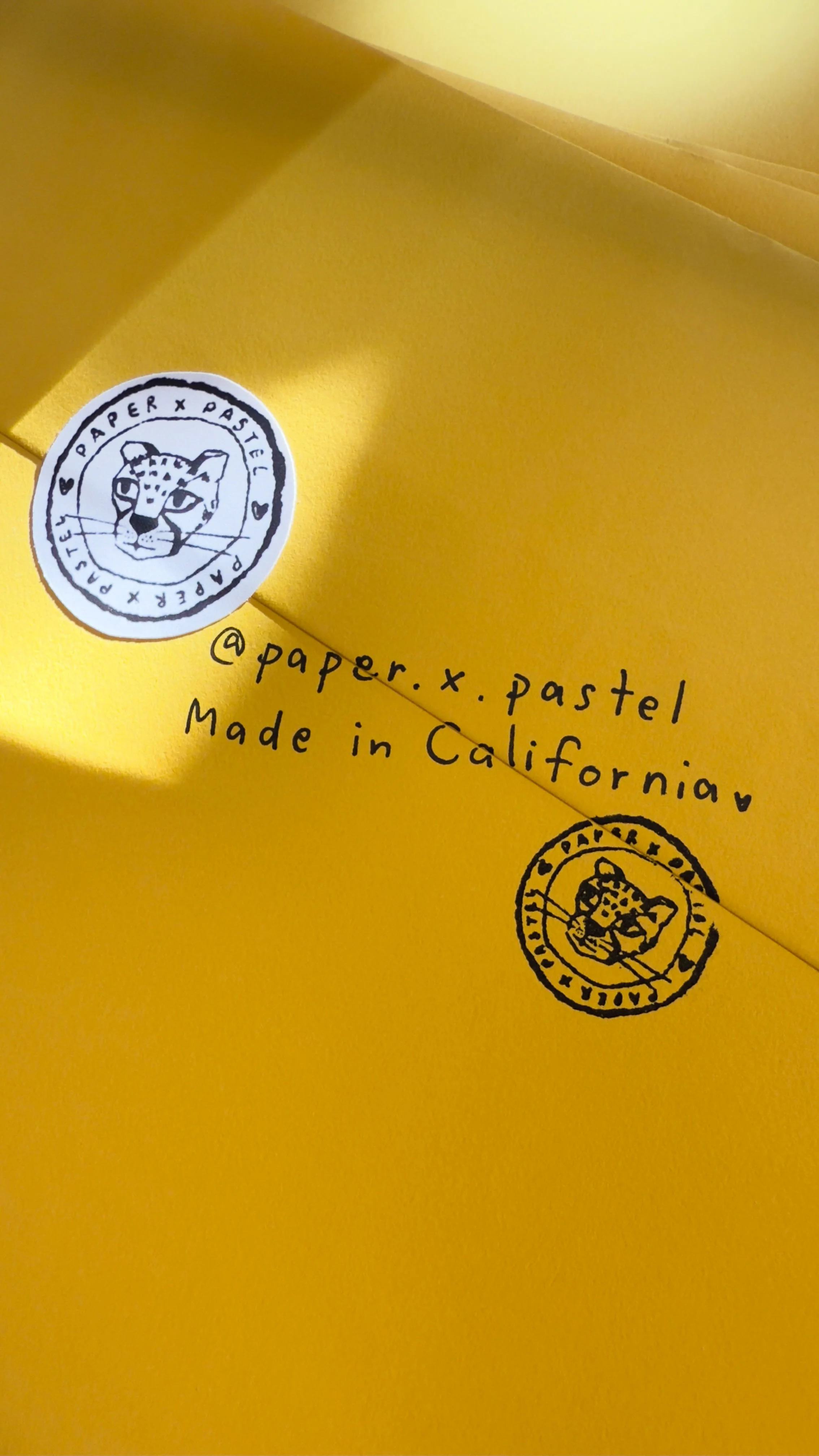 Close-up of yellow packaging or paper with black handwritten text '@paper.x.pastel Made in California' and two circular stamps featuring a leopard's head, one in black and one in white.