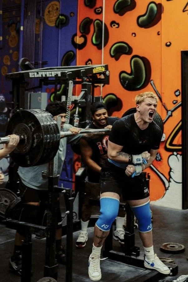 A woman in a black shirt and shorts is doing a squat with weights on her shoulders, while three men cheer her on in a colorful gym with climbing wall art in the background.
