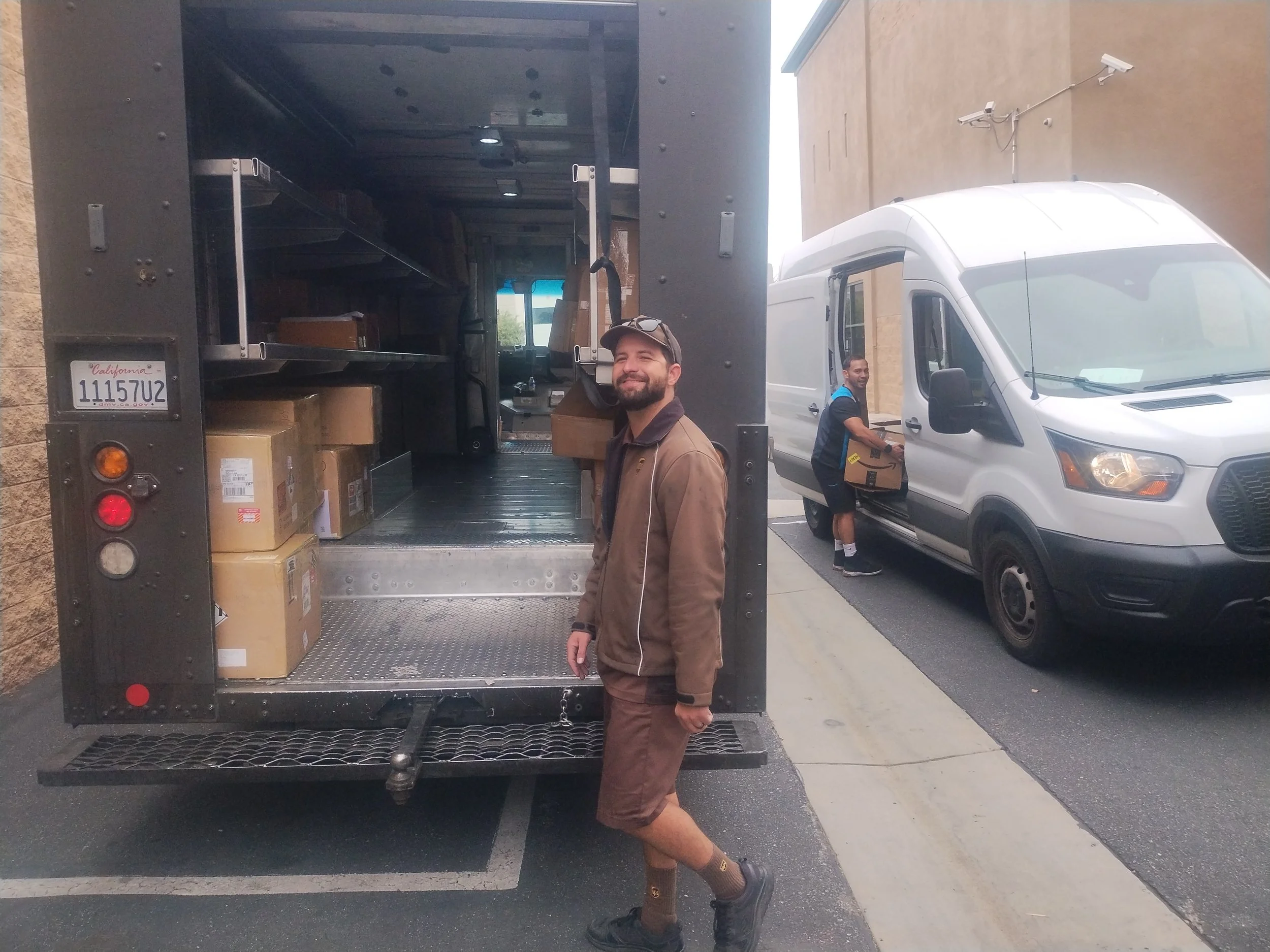 Two delivery men loading boxes into a white delivery truck parked in a parking lot next to a building. One man is standing outside the truck smiling, while the other is loading boxes into the truck.